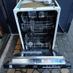 Sebastian Warchoł - Otwarta, uszkodzona zmywarka AEG na zewnątrz, widoczne wnętrze i elementy instalacji. Sprzęt stoi na kostce brukowej, tło stanowi ciemna ściana. Widoczne ślady użytkowania.