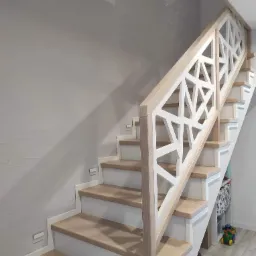 Jasne, drewniane schody z białą, geometryczną balustradą, oświetleniem punktowym w ścianie i zabawkami dziecięcymi w tle.