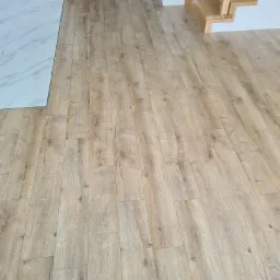 Ułożone panele podłogowe w jasnym odcieniu drewna, widoczne schody z naturalnego drewna i fragment marmurowej posadzki. Nowoczesne wykończenie wnętrza.