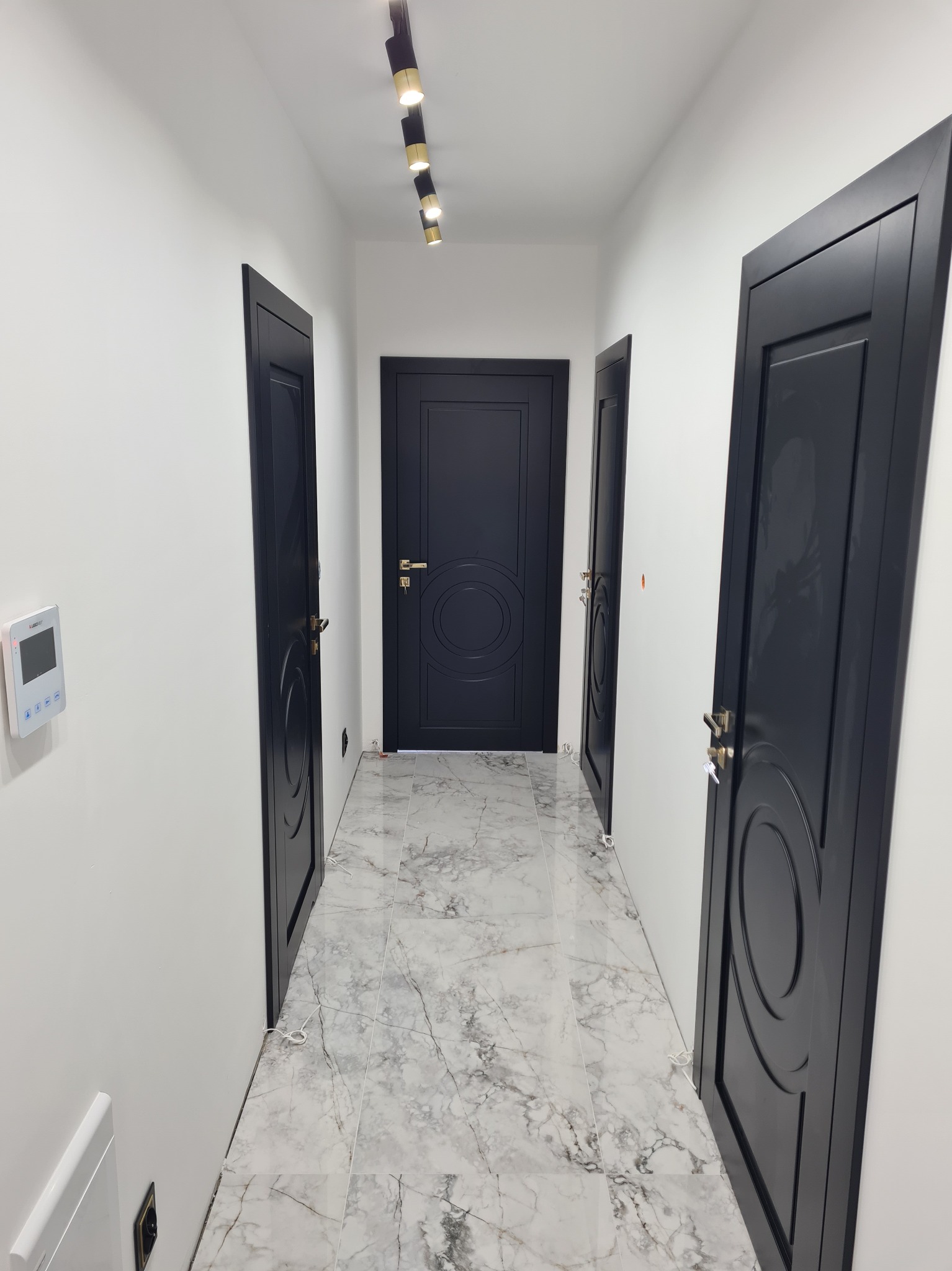 Nowoczesny korytarz z trzema czarnymi drzwiami wewnętrznymi z dekoracyjnymi okręgami, oświetlony listwą LED. Marmurowe płytki podłogowe. Minimalistyczny design.