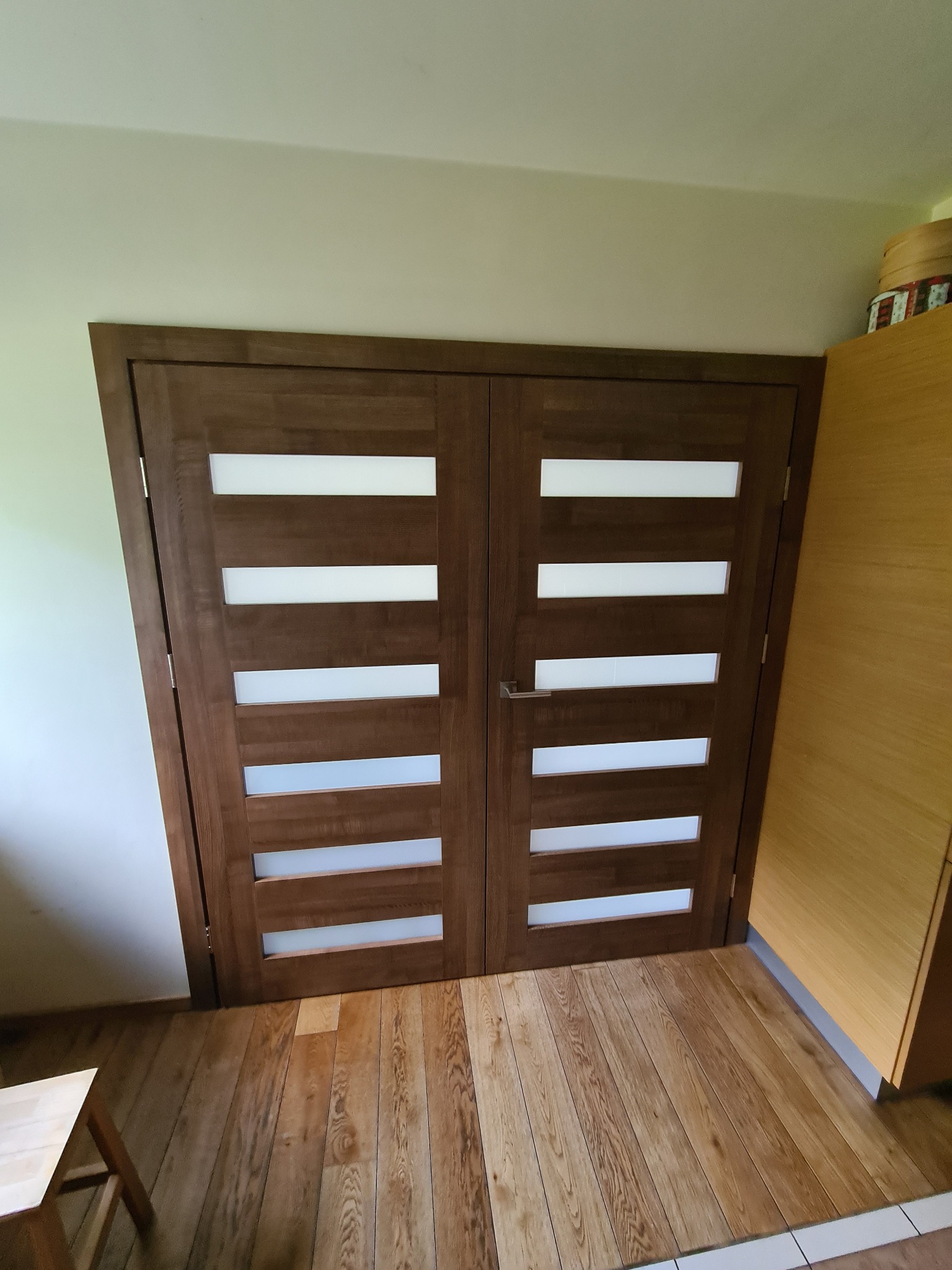 Dwuskrzydłowe, brązowe drzwi wewnętrzne z poziomymi, mlecznymi przeszkleniami, klamka po prawej stronie, widok z bliska na parkiet, fragment taboretu po lewej.