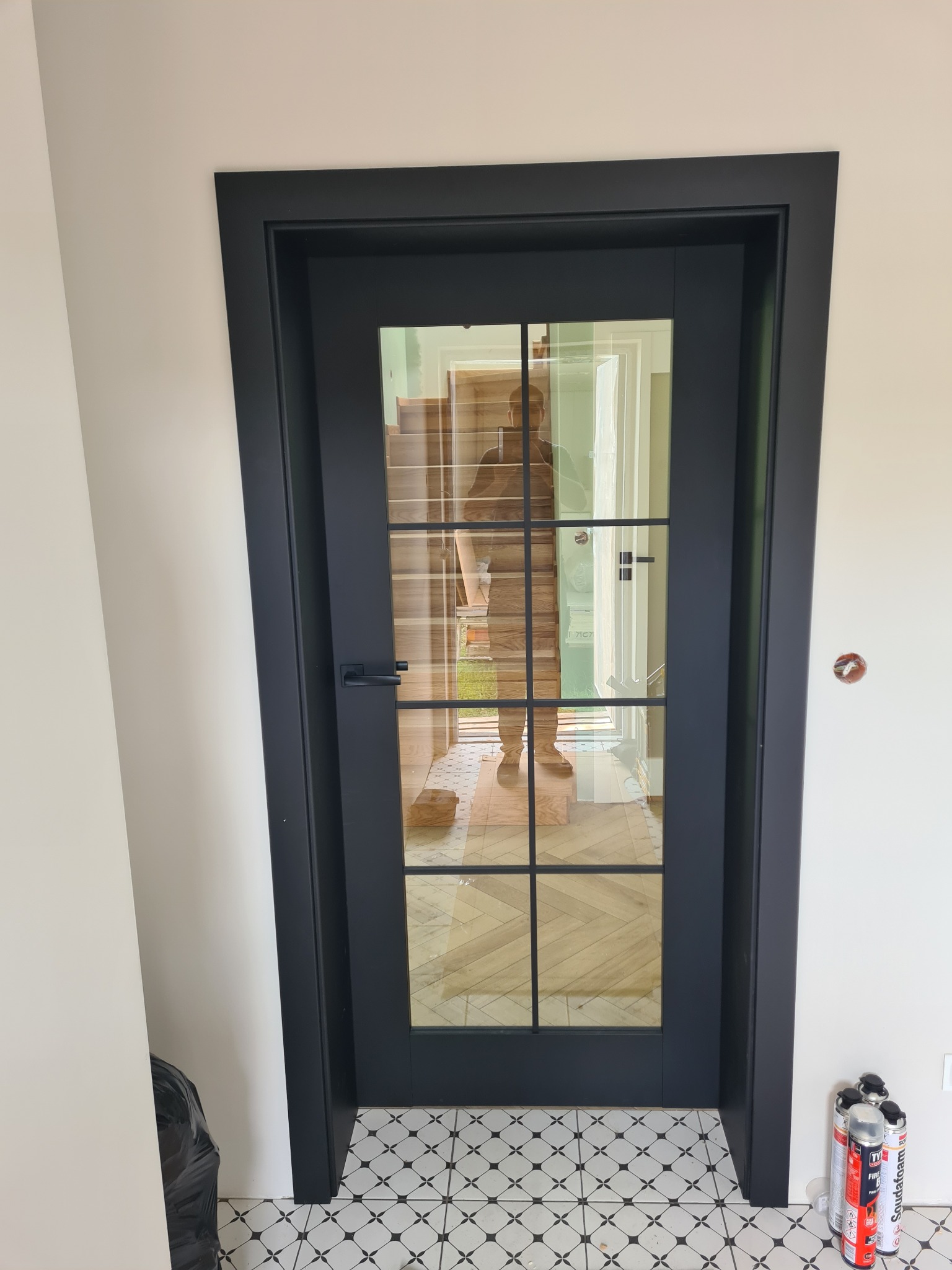 Czarna, przeszklona rama drzwi wewnętrznych z widocznym odbiciem osoby, osadzona w białej ścianie nad wzorzystą podłogą. Widoczne puszki z pianką montażową.