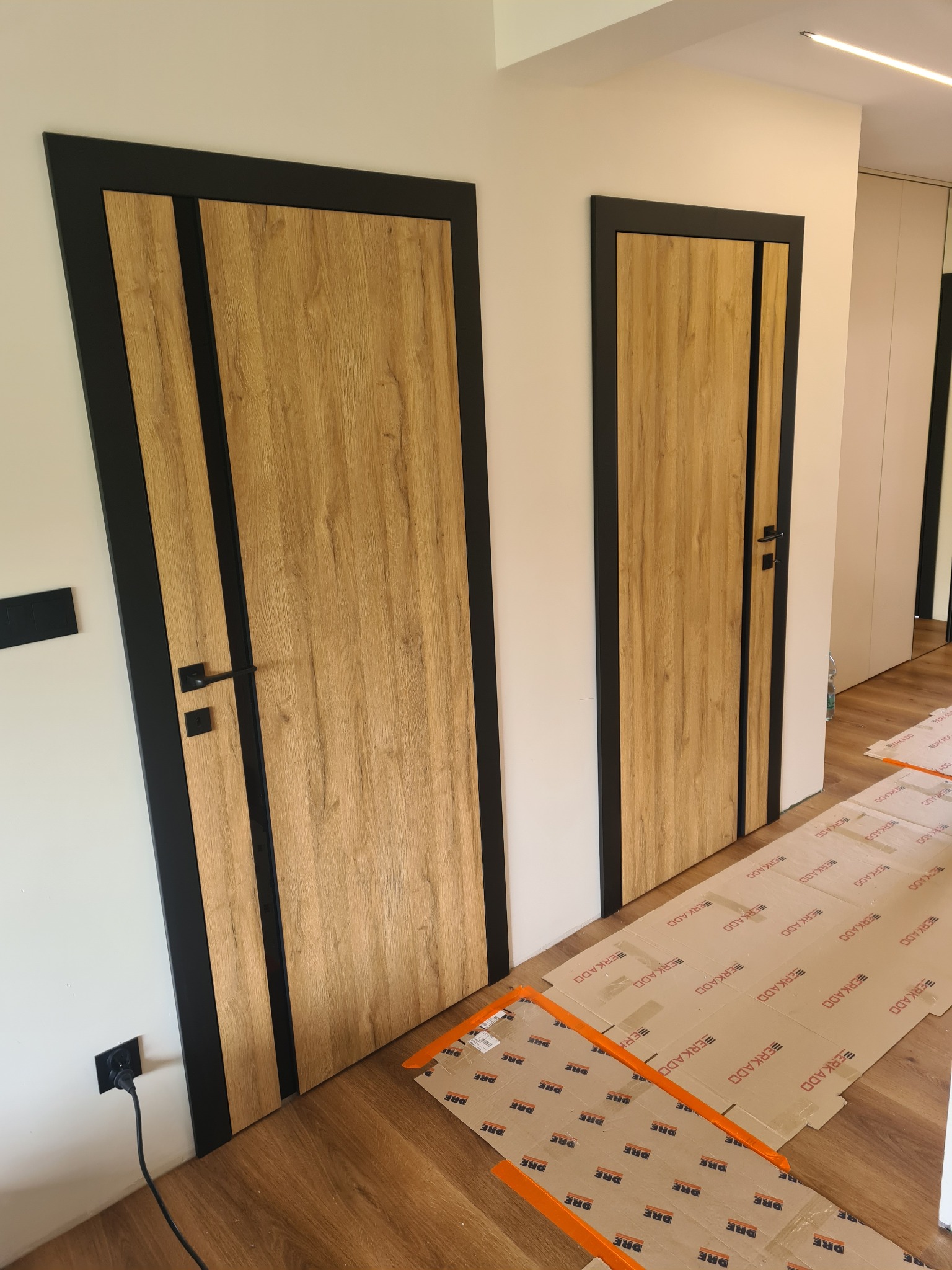 Dwa zamontowane, drewnopodobne drzwi wewnętrzne z czarną ramą i klamką. Na podłodze kartony zabezpieczające. Widok na korytarz i szafę wnękową w tle.