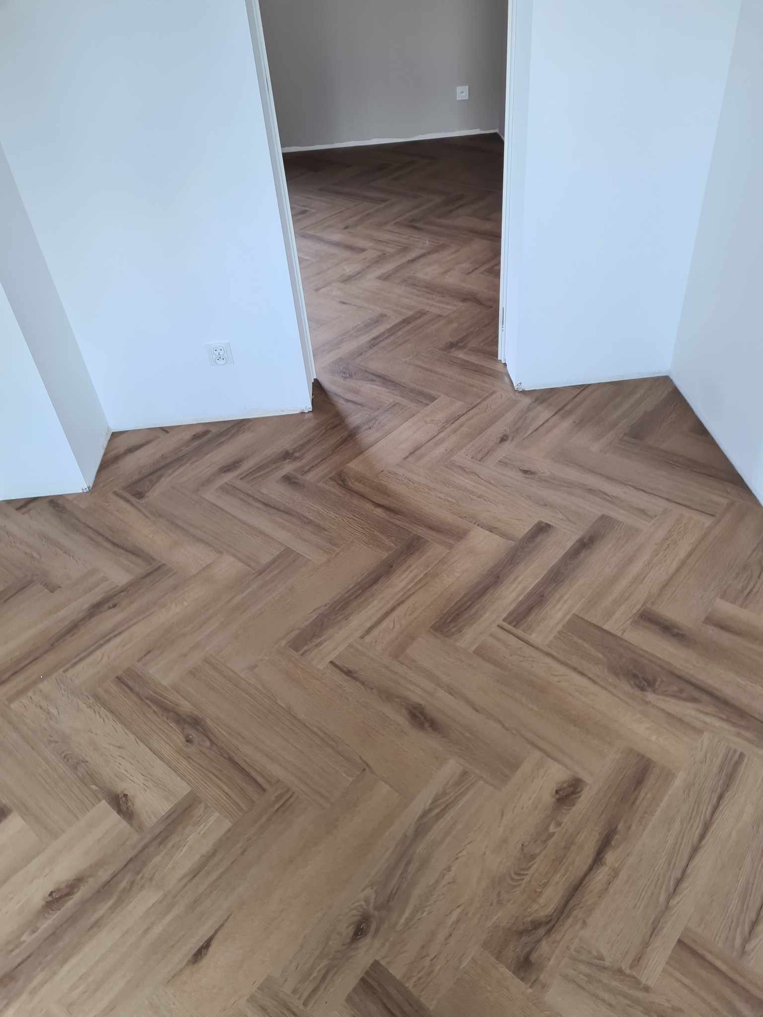 Parkiet ułożony we wzór jodełki w jasnym, nowym wnętrzu. Widoczny fragment białej ściany z gniazdkiem i przejście do kolejnego pomieszczenia z identyczną podłogą.