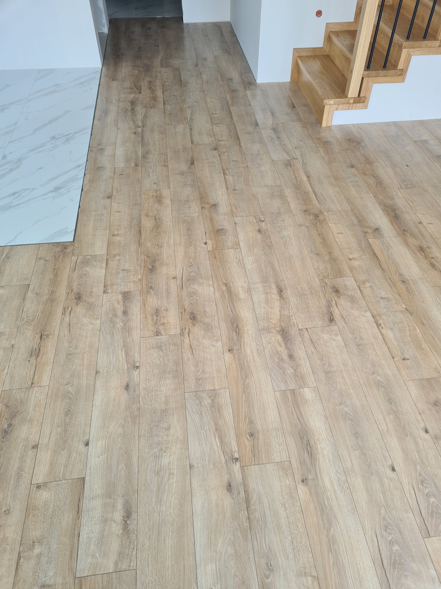 Ułożone panele podłogowe w jasnym odcieniu drewna, widoczne schody z naturalnego drewna i fragment marmurowej posadzki. Nowoczesne wykończenie wnętrza.