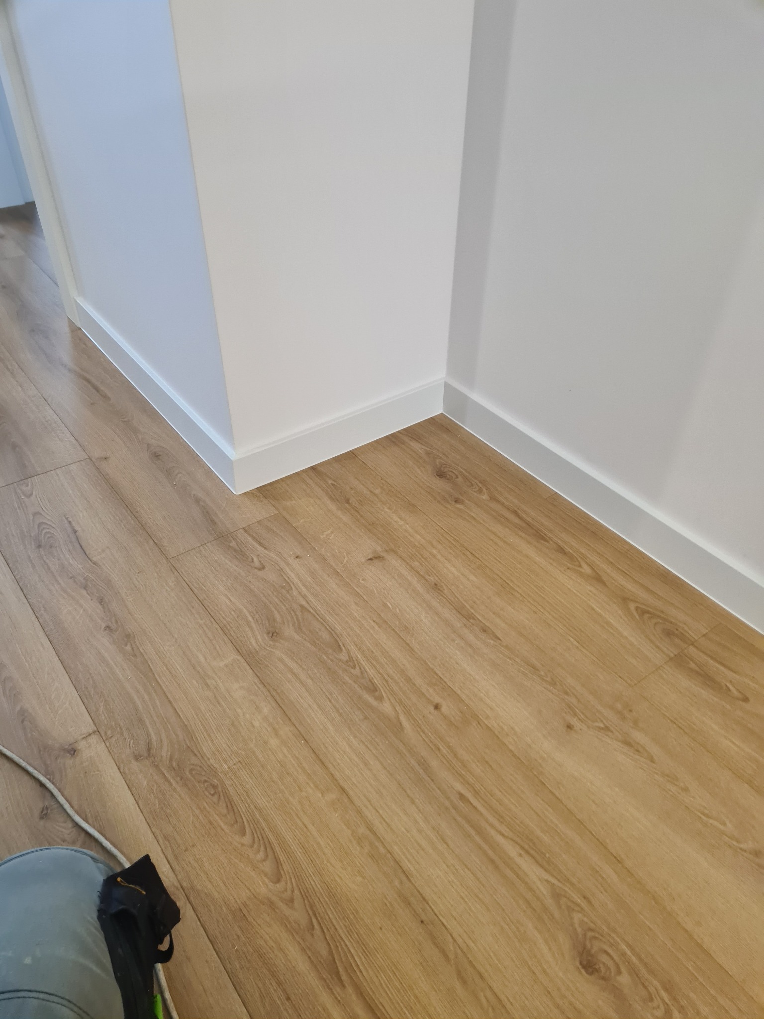 Jasne panele podłogowe ułożone w rogu pomieszczenia z białymi listwami przypodłogowymi, widok z góry, fragment buta roboczego w lewym dolnym rogu.