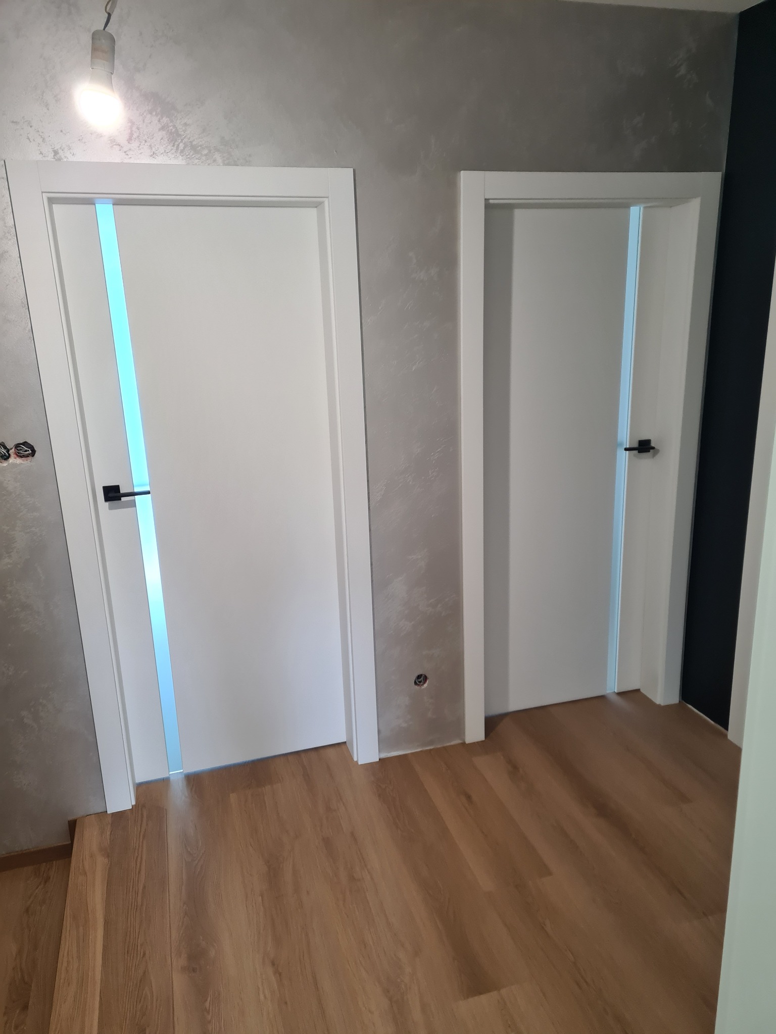 Dwa białe skrzydła drzwiowe z czarnymi klamkami w nowym budownictwie. Jedne ze szklanym panelem. Na ścianie widoczne gniazdko elektryczne przed wykończeniem.