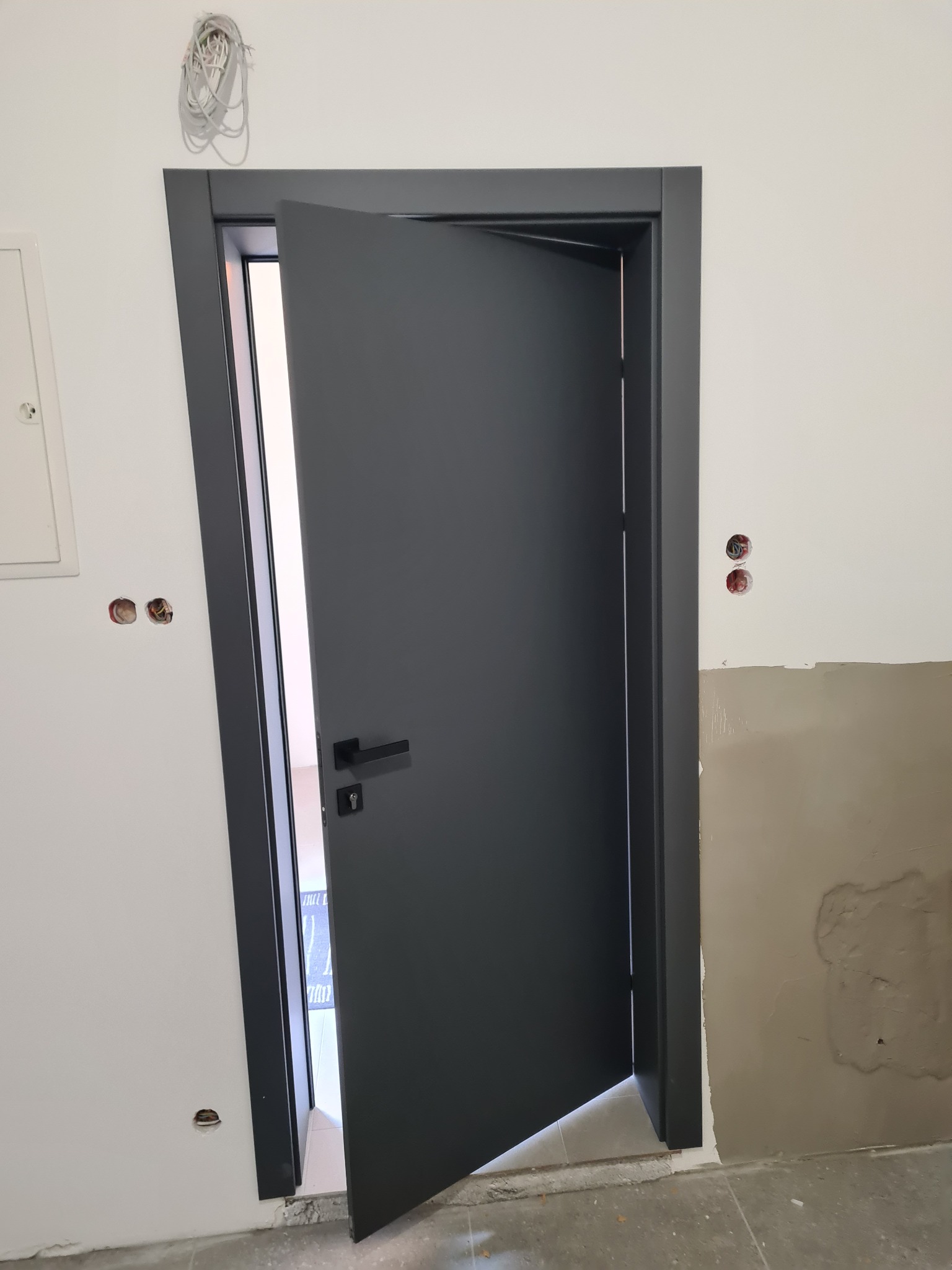 Ciemnoszare, częściowo otwarte drzwi wewnętrzne z czarną klamką, osadzone w szarej futrynie. Widoczne niedokończone ściany z instalacją elektryczną.