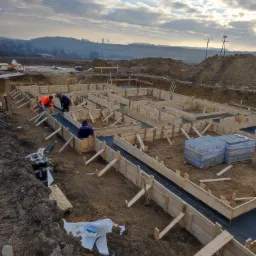 Przygotowanie szalunku i zbrojenia pod fundament domu jednorodzinnego na działce z widokiem na pagórkowaty krajobraz w pochmurny dzień.