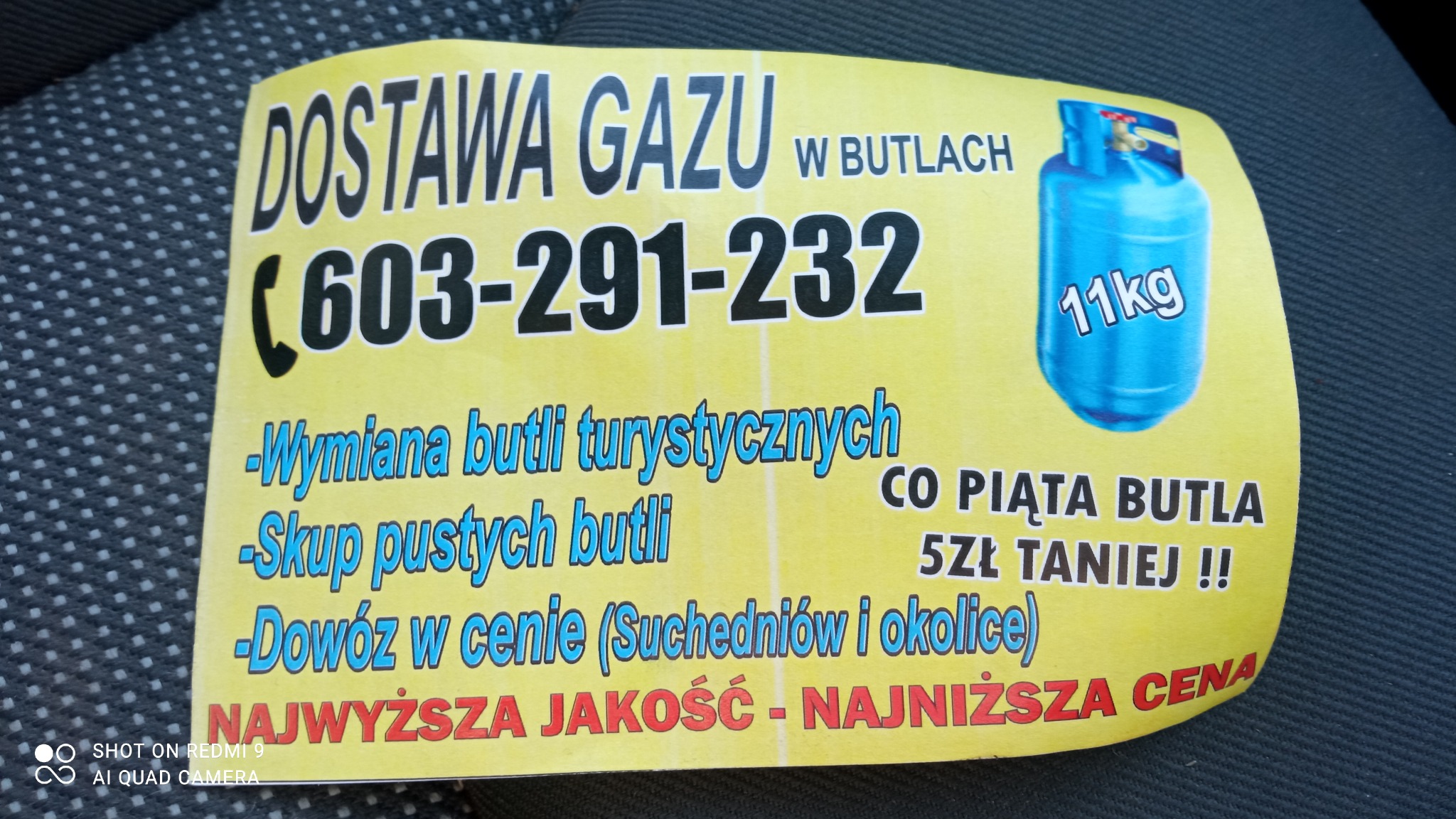 Żółta ulotka reklamowa z informacją o dostawie gazu w butlach, numerem telefonu i hasłem 'Najwyższa jakość - najniższa cena', leżąca na ciemnej tapicerce.