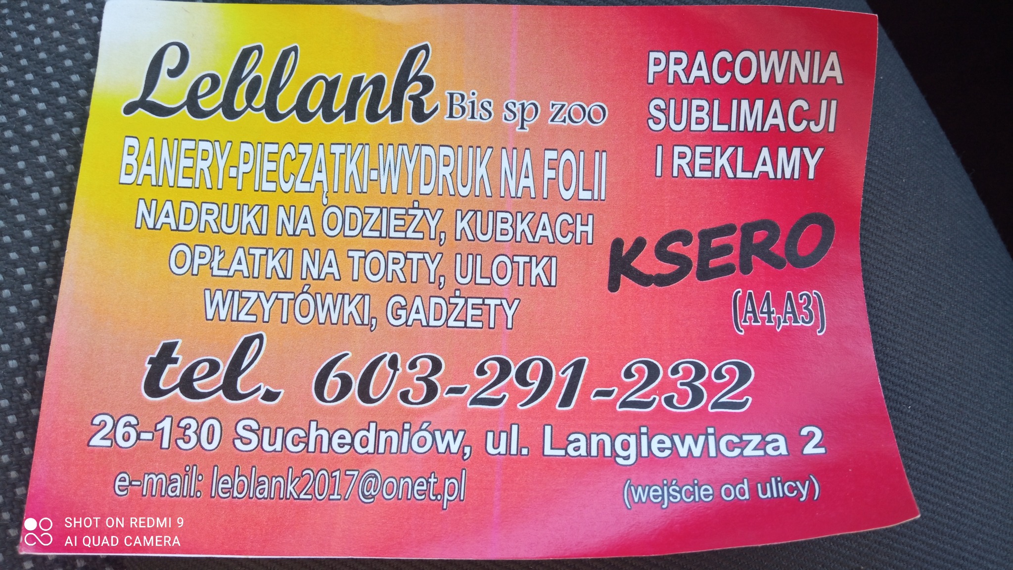 Żółto-czerwona ulotka reklamowa firmy Leblank z Suchedniowa, oferującej nadruki na odzieży, banery, wizytówki, ulotki i ksero, leżąca na ciemnej, tekstylnej powierzchni.