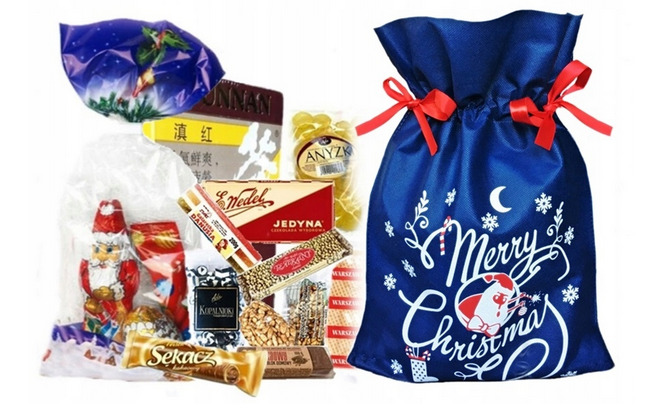 Różnorodność słodyczy i przekąsek, w tym czekoladowy Mikołaj, wafle i batony, zapakowane w przezroczyste folie i ozdobny, granatowy worek ze świątecznym nadrukiem Merry Christmas, zawiązany...