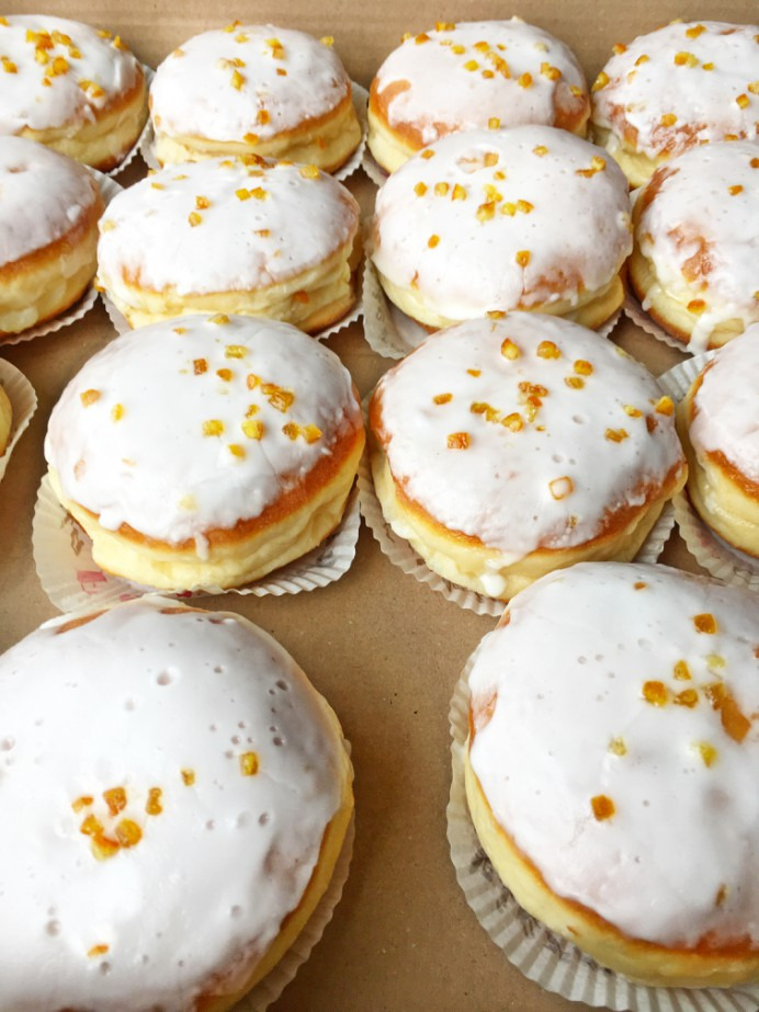 Słodkie pączki z białym lukrem i kandyzowaną skórką pomarańczową, ułożone w kartonowym pudełku na papierowych podkładkach.