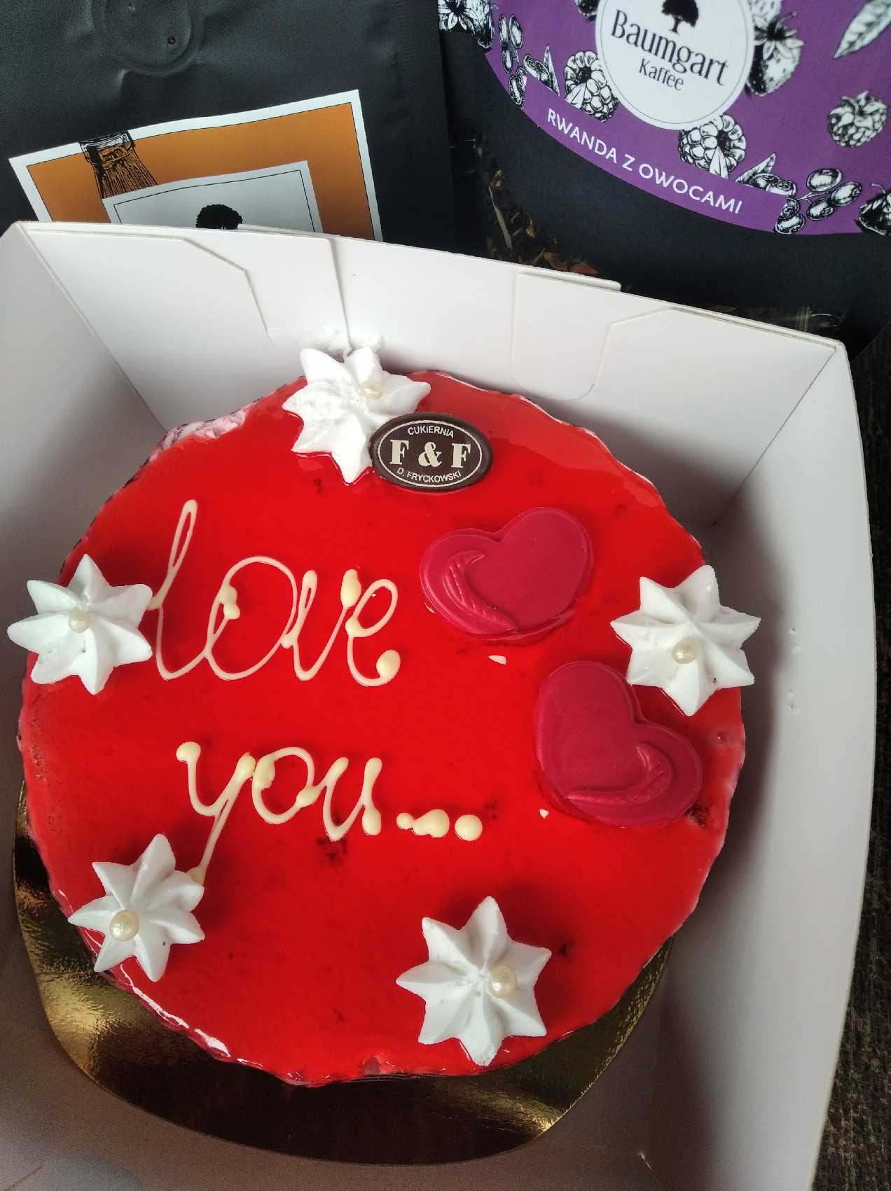 Czerwony tort z napisem 'love you...', dekorowany białymi gwiazdkami z perłami i dwoma czerwonymi sercami, umieszczony w białym pudełku obok kawy Baumgart Rwanda z owocami i kartki.