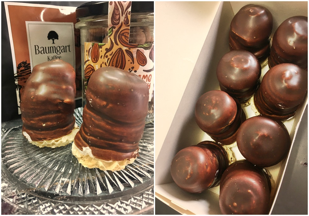 Dwa czekoladowe pianki na waflowym spodzie, prezentowane na szklanym talerzu z wzorem, obok słoika z orzechami i opakowania kawy Baumgart Kaffee; pudełko z piankami w tle.