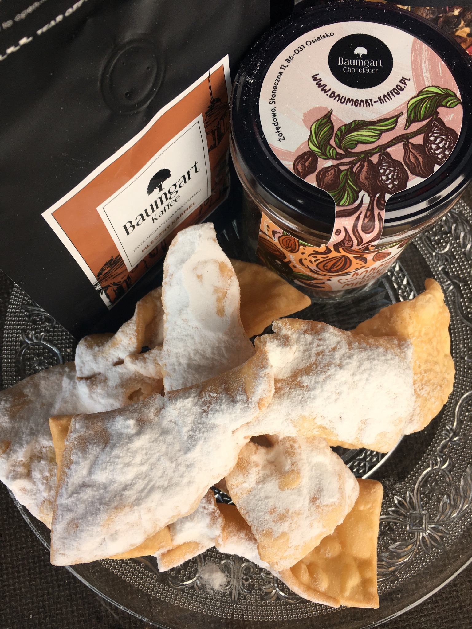 Faworki posypane cukrem pudrem na szklanym talerzu, obok kawa i czekolada Baumgart, widoczne opakowania z logo firmy.