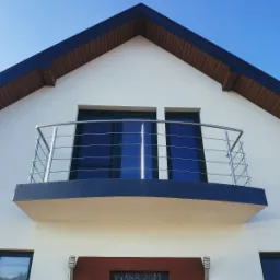 Balkon z metalową konstrukcją zabezpieczającą na tle błękitnego nieba, widok z dołu na białą fasadę domu z ciemnymi oknami.