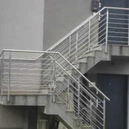 Zewnętrzne schody z szarego granitu z nowoczesną, metalową barierką z poziomych prętów, prowadzące do dwóch wejść w budynku mieszkalnym.