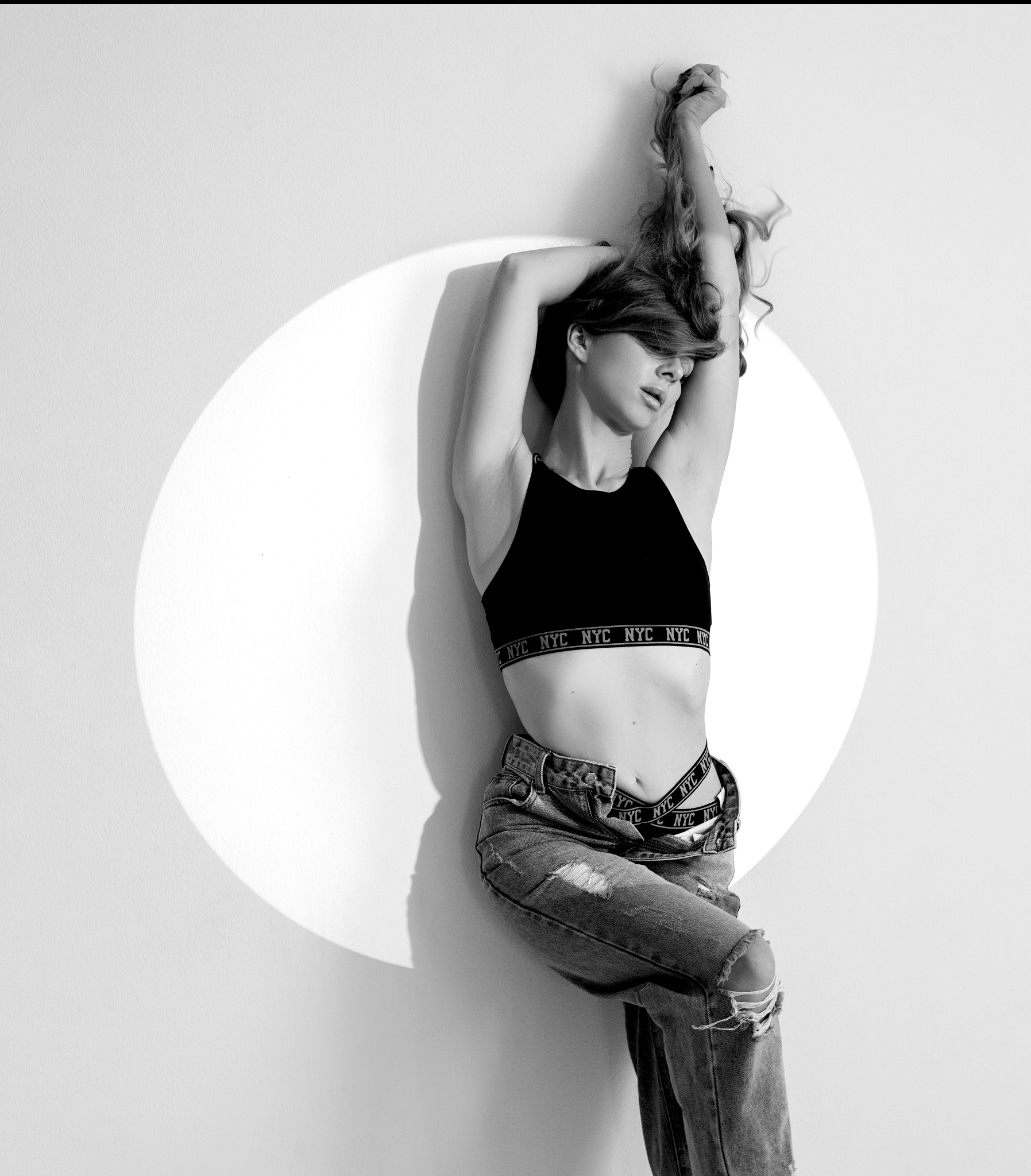 Czarno-białe zdjęcie kobiety w sportowym topie i podartych jeansach, opierającej się o ścianę z okrągłym światłem padającym na nią.