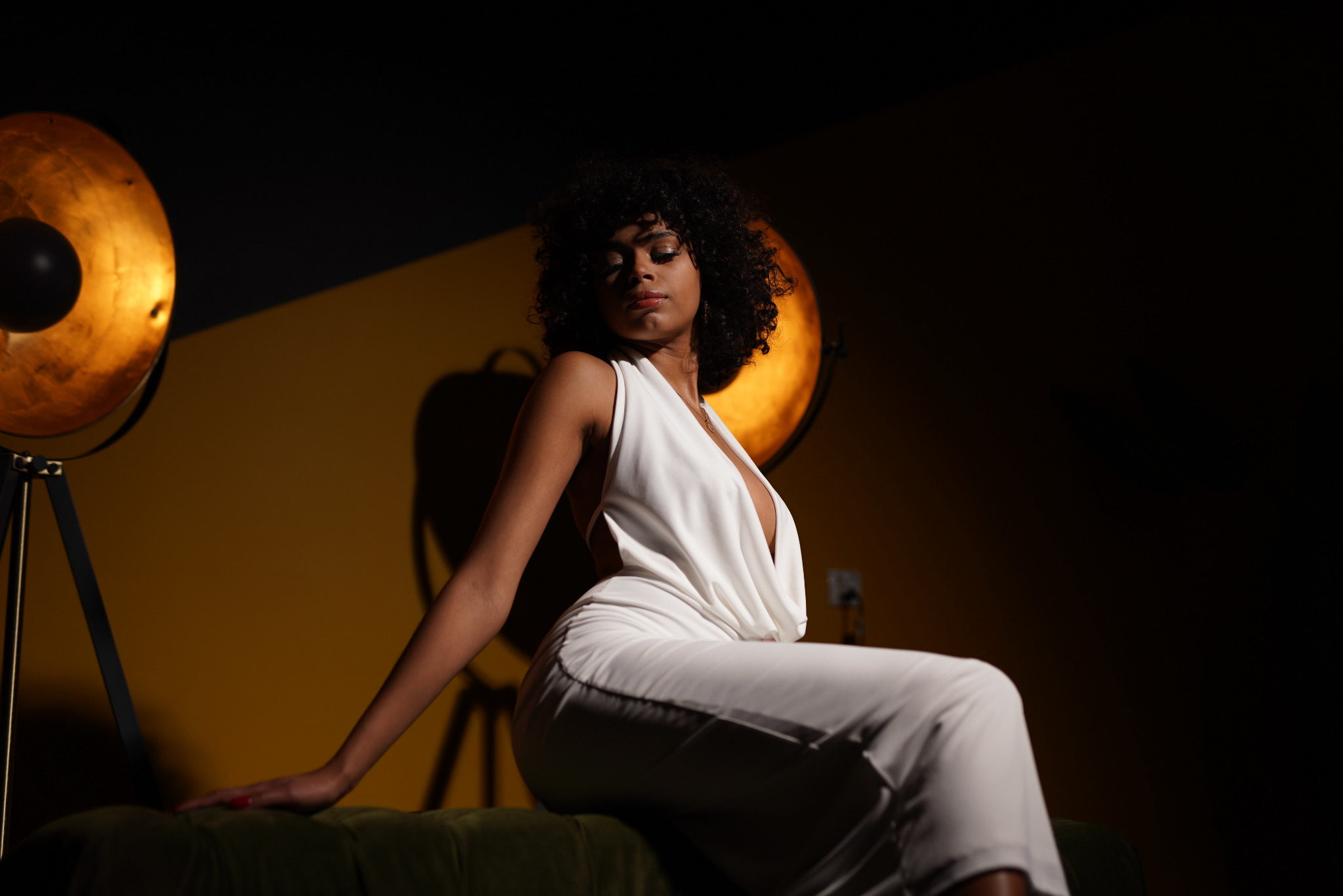 Portret kobiety w białym kombinezonie, siedzącej na zielonej sofie, oświetlonej przez lampę studyjną z żółtym tłem.