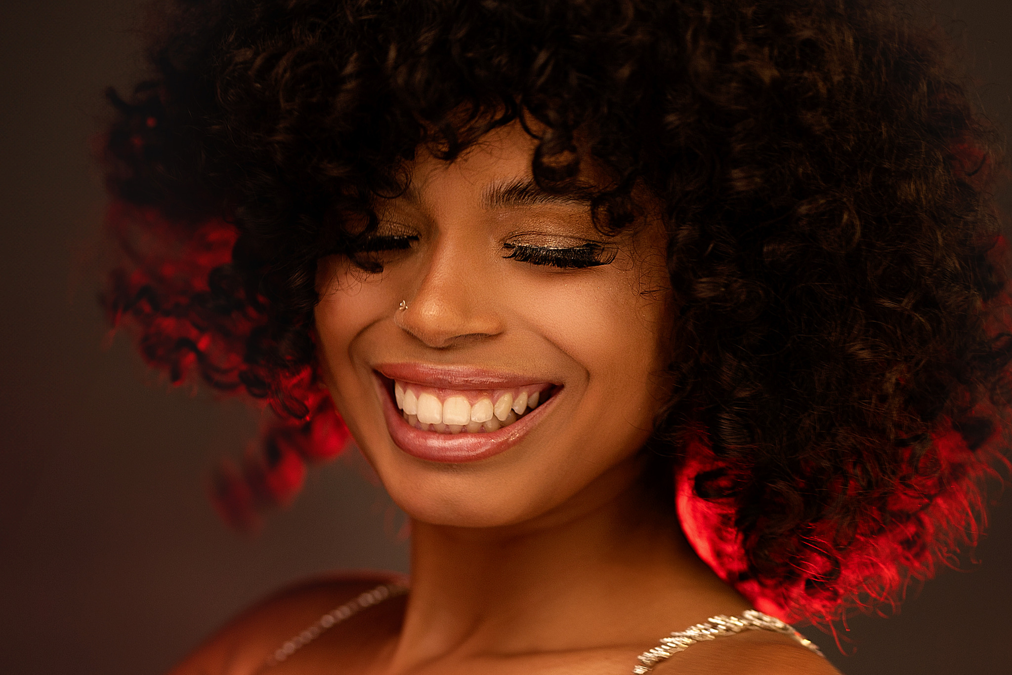 Portret uśmiechniętej młodej kobiety z afro, złoty makijaż oczu i kolczyk w nosie, oświetlona czerwonym światłem od tyłu.