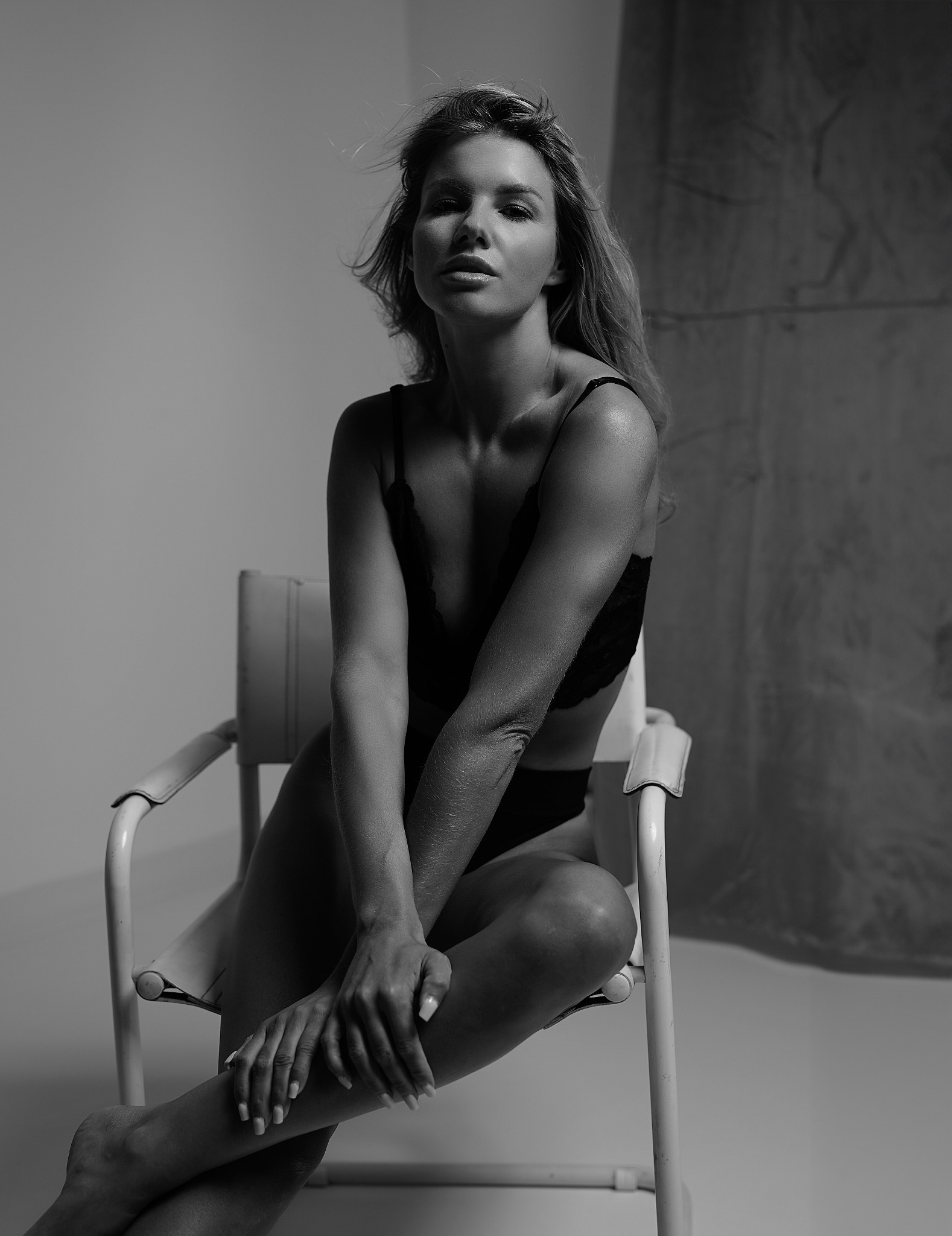 Czarno-białe portretowe ujęcie kobiety siedzącej na krześle w bieliźnie, z delikatnym wiatrem we włosach i skupionym spojrzeniem.