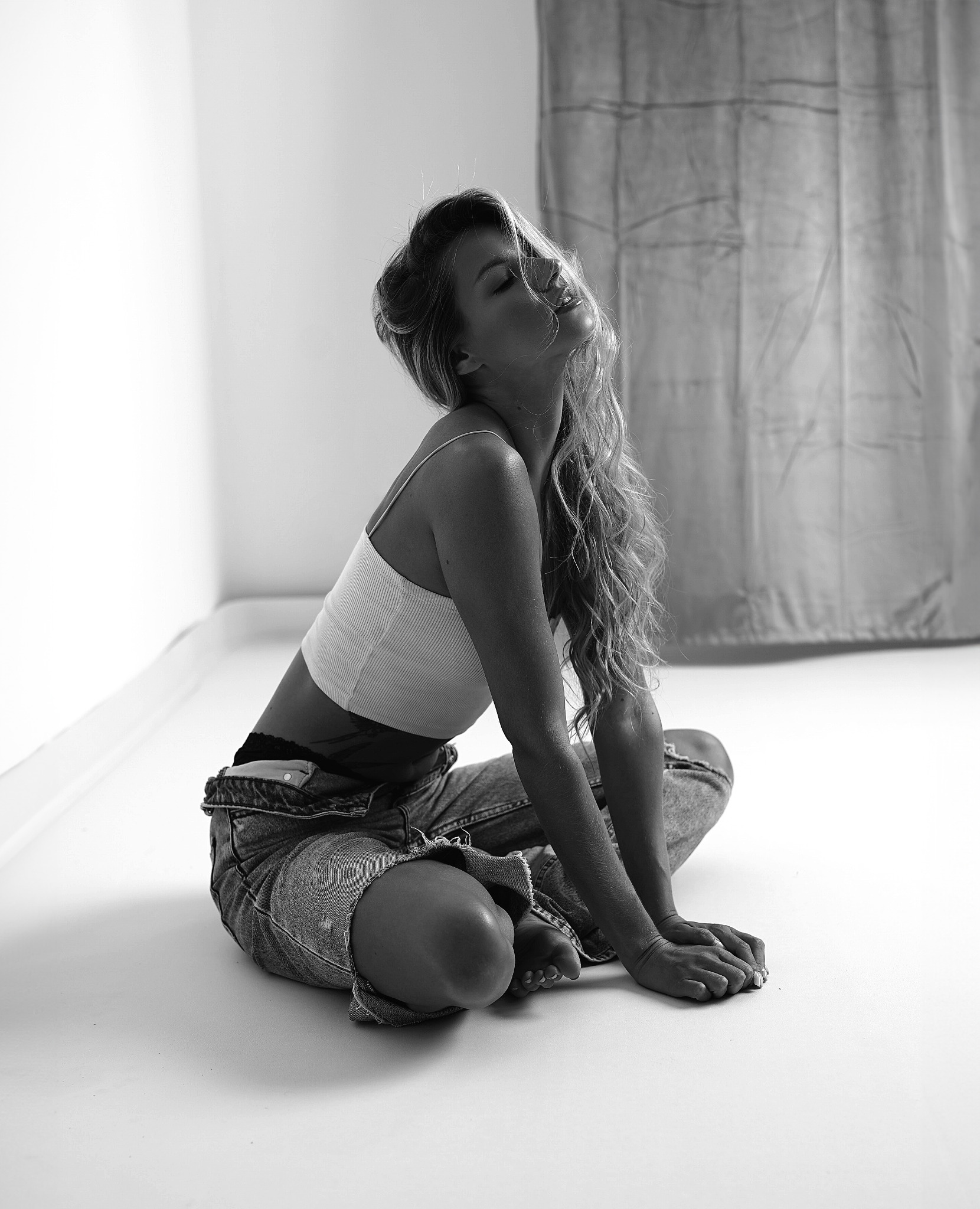 Czarno-białe zdjęcie kobiety siedzącej na podłodze w dżinsowych ogrodniczkach i krótkim topie, z zamkniętymi oczami i głową odchyloną do tyłu, w minimalistycznym wnętrzu z zasłoną w tle.