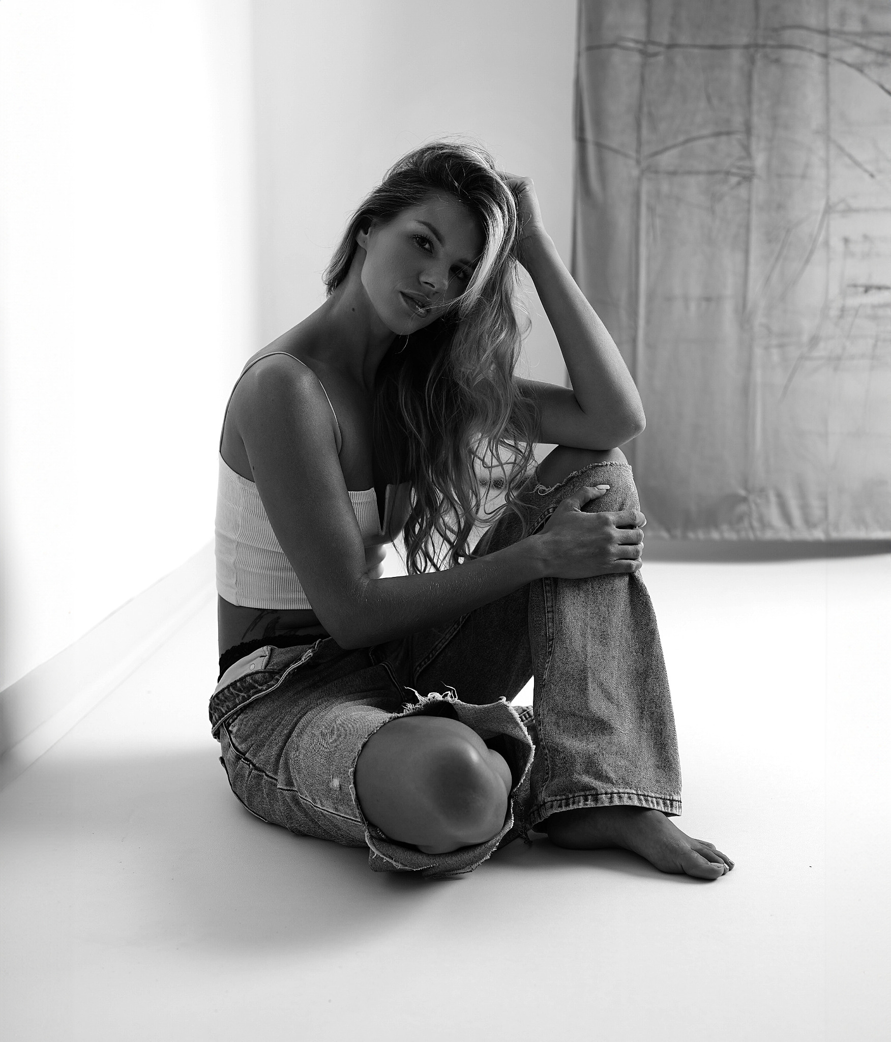 Portret kobiety siedzącej na podłodze w podartych jeansach i topie, z ręką we włosach, w czarno-białej tonacji, na tle jasnej ściany i abstrakcyjnej tkaniny.