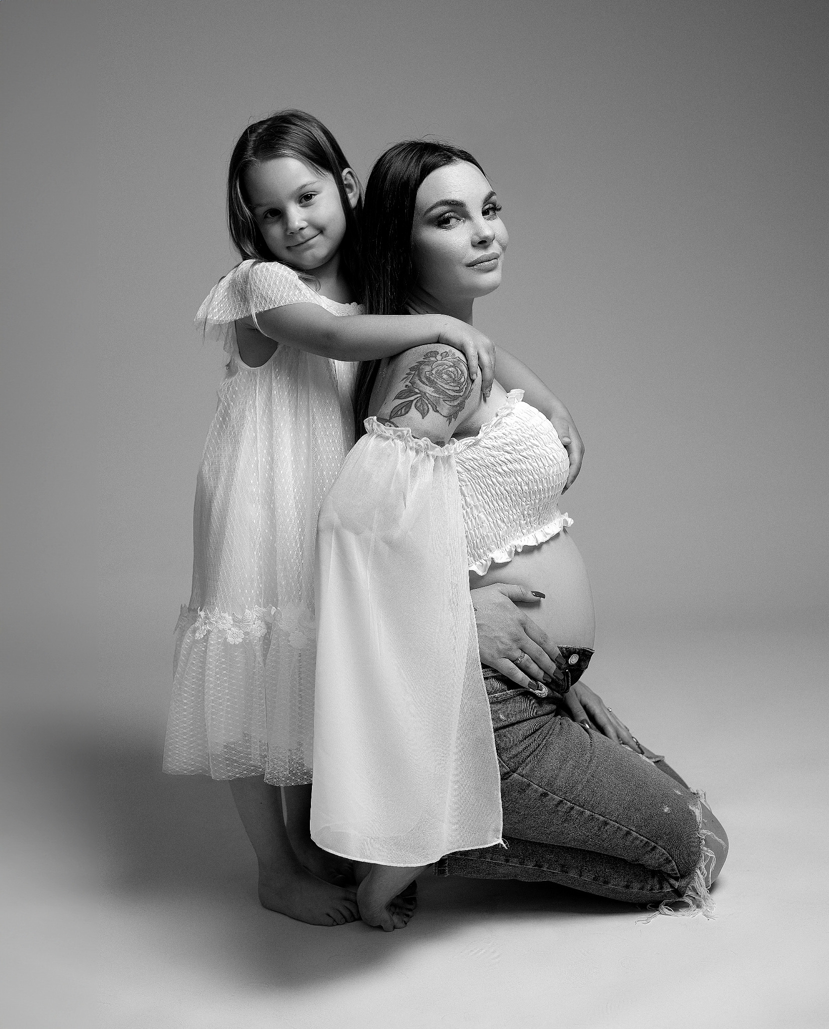 Czarno-białe zdjęcie ciężarnej kobiety klęczącej na podłodze, obejmowanej przez córkę ubraną w białą sukienkę, z widocznym tatuażem róży na ramieniu kobiety.