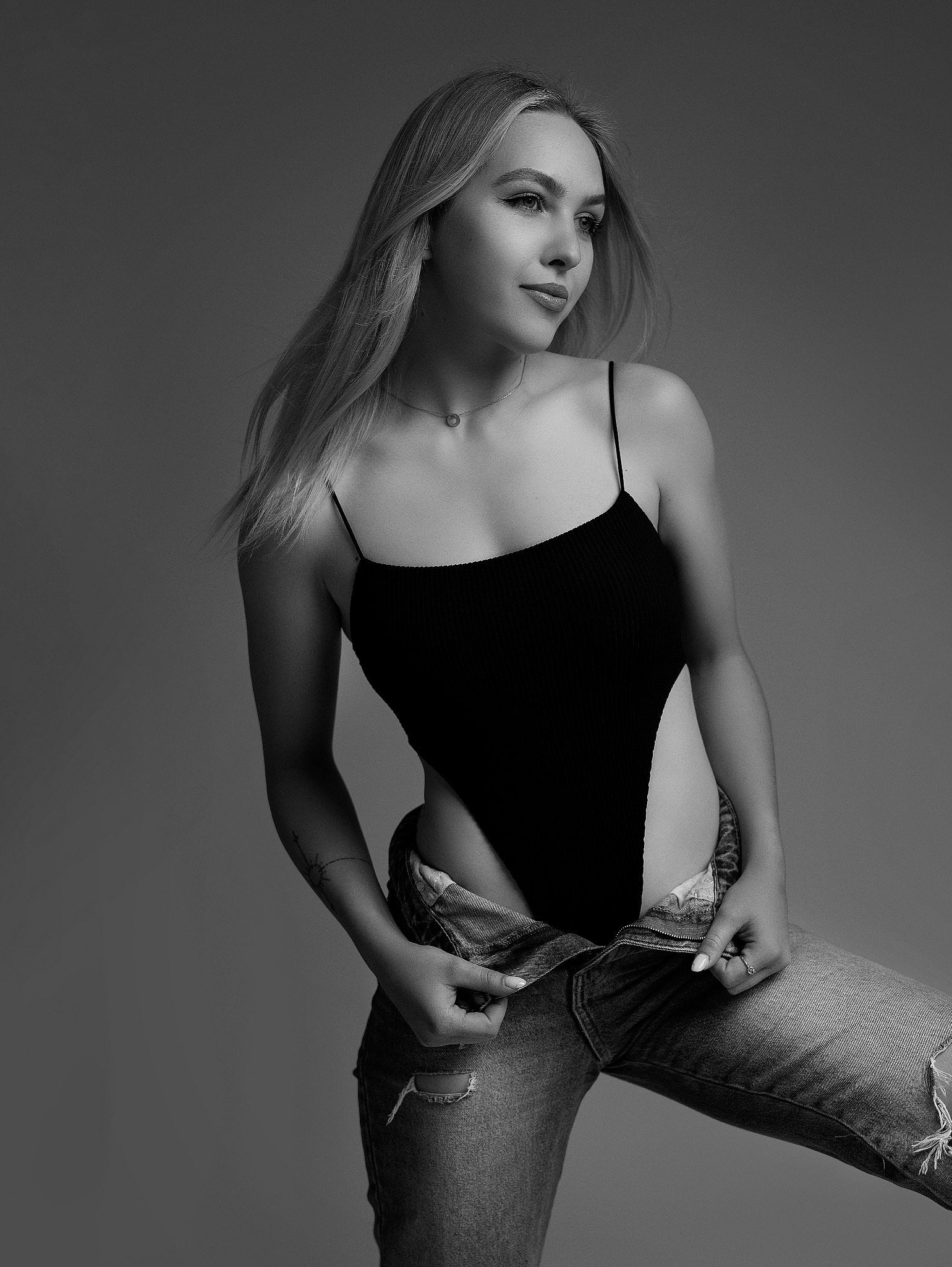 Czarno-białe portretowe ujęcie kobiety z rozwianymi blond włosami, w czarnym body na ramiączkach i rozpiętych dżinsach z dziurami, spoglądającej w lewo.