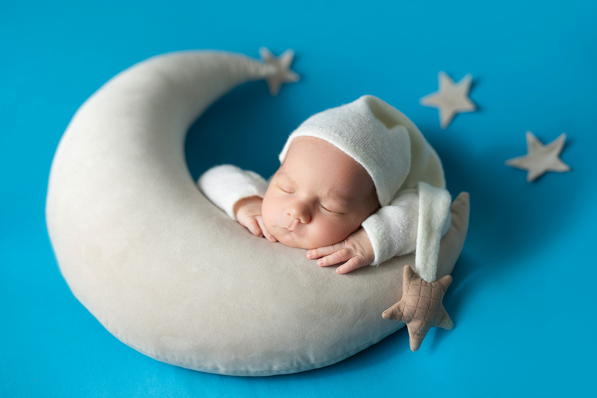 Noworodek śpiący na poduszce w kształcie księżyca, w białym ubranku i czapeczce, otoczony pluszowymi gwiazdkami na błękitnym tle.