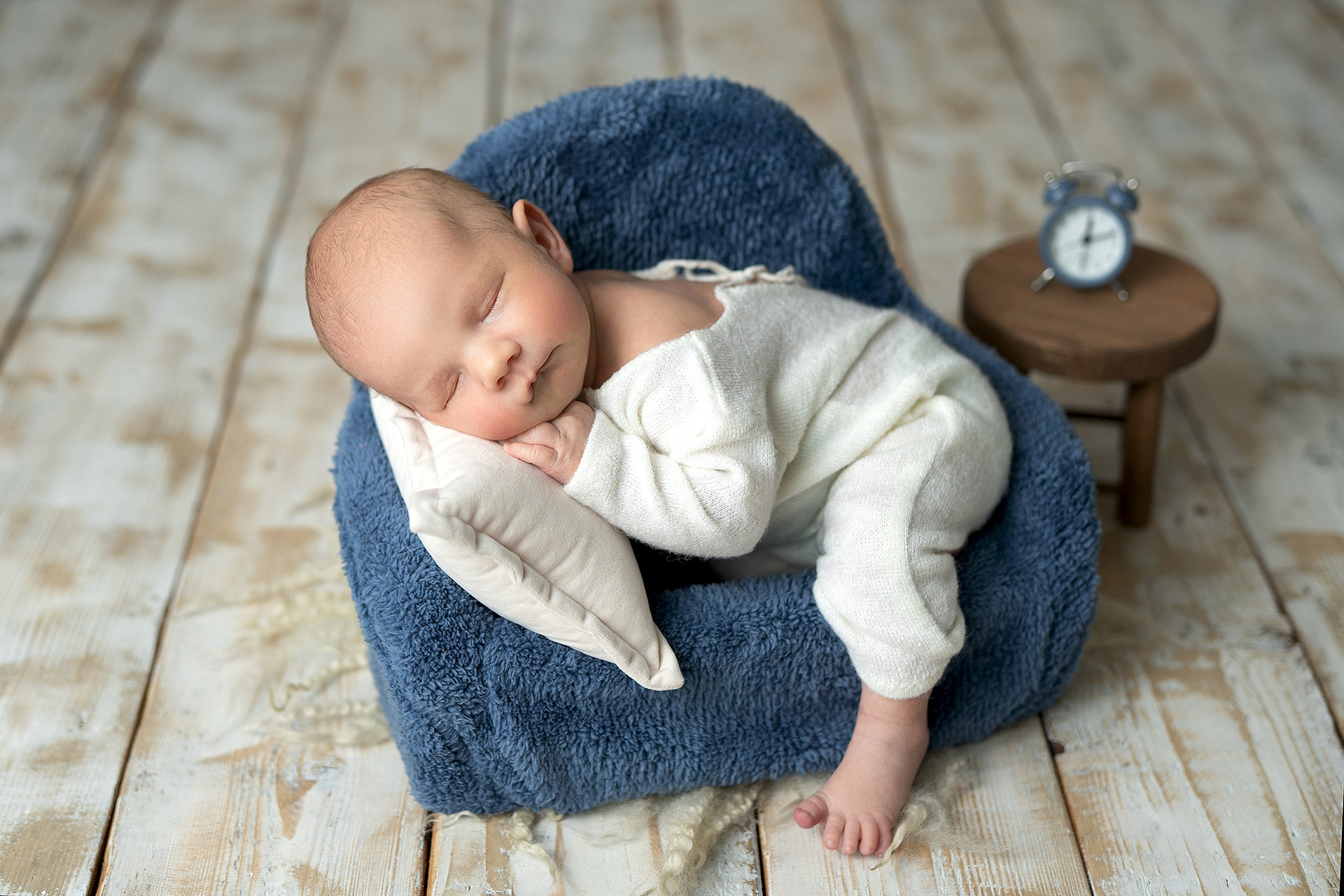 Noworodek śpiący w niebieskim fotelu na drewnianej podłodze, obok mały stolik z budzikiem