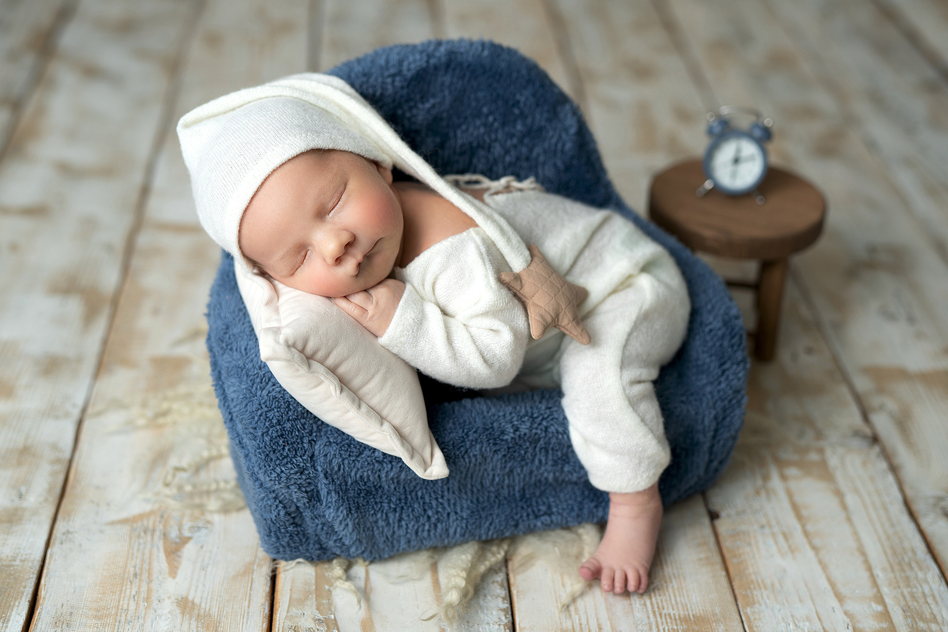 Noworodek w białym stroju i czapce śpi na niebieskim pluszowym fotelu z poduszką w studio w Tarnowie, obok stoi mały drewniany stołek z budzikiem.