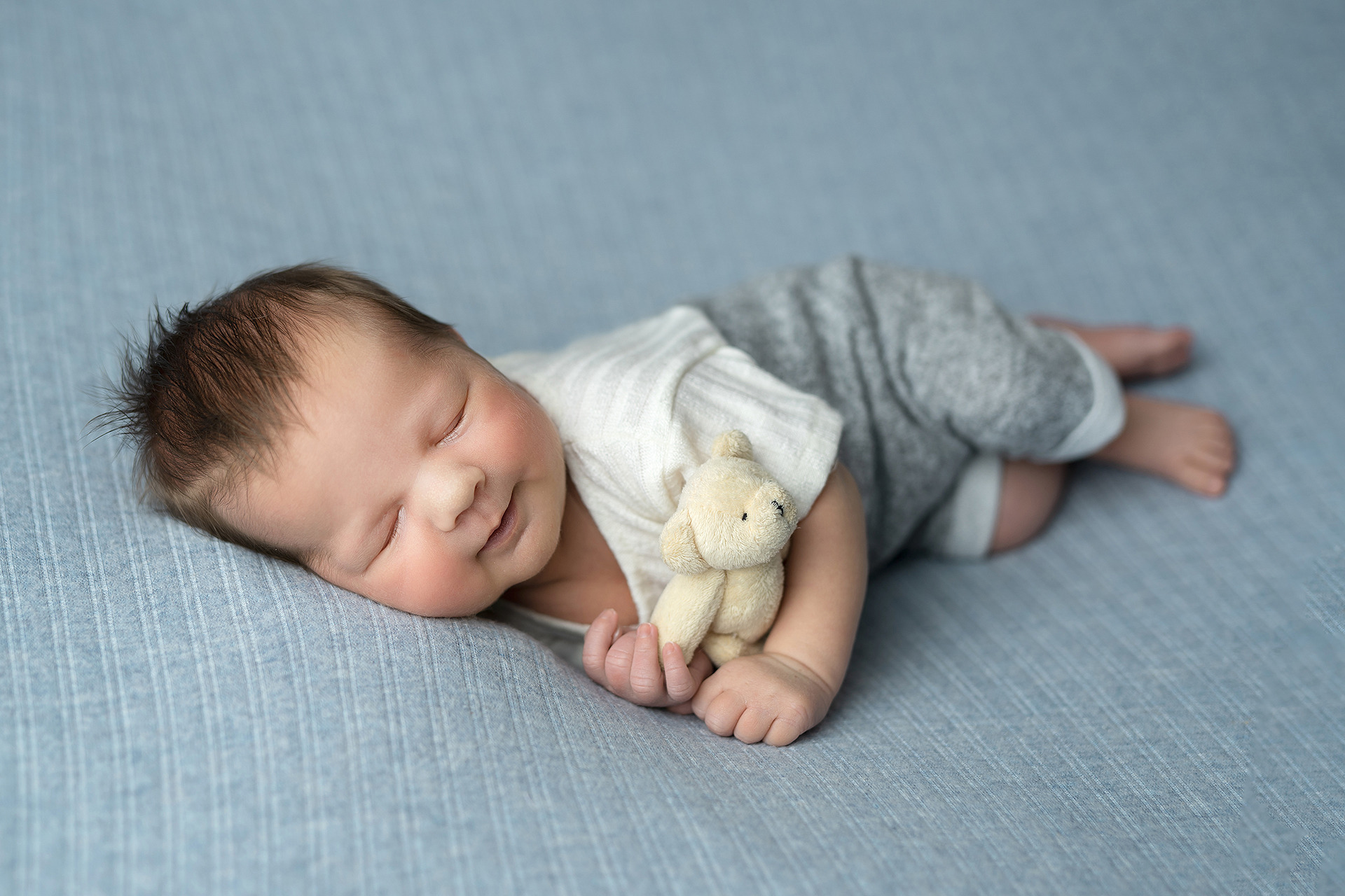 Śpiący noworodek w szarym ubranku, trzymający małego pluszowego misia na błękitnym tle.