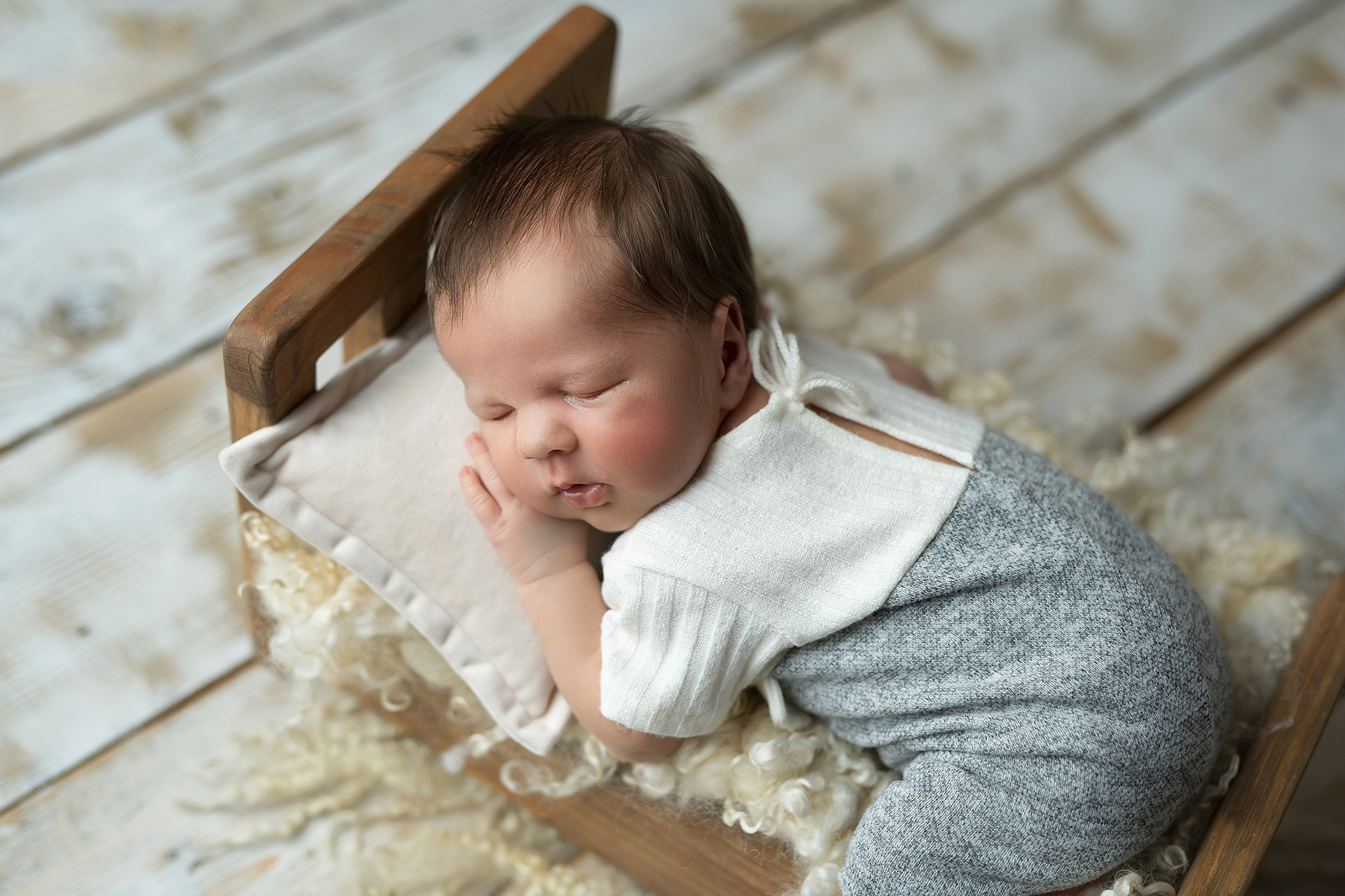 Noworodek śpiący w małym drewnianym łóżeczku na miękkim, kremowym futerku, ubrany w szaro-biały strój z wiązanymi sznureczkami przy szyi, dłoń podłożona pod policzek.