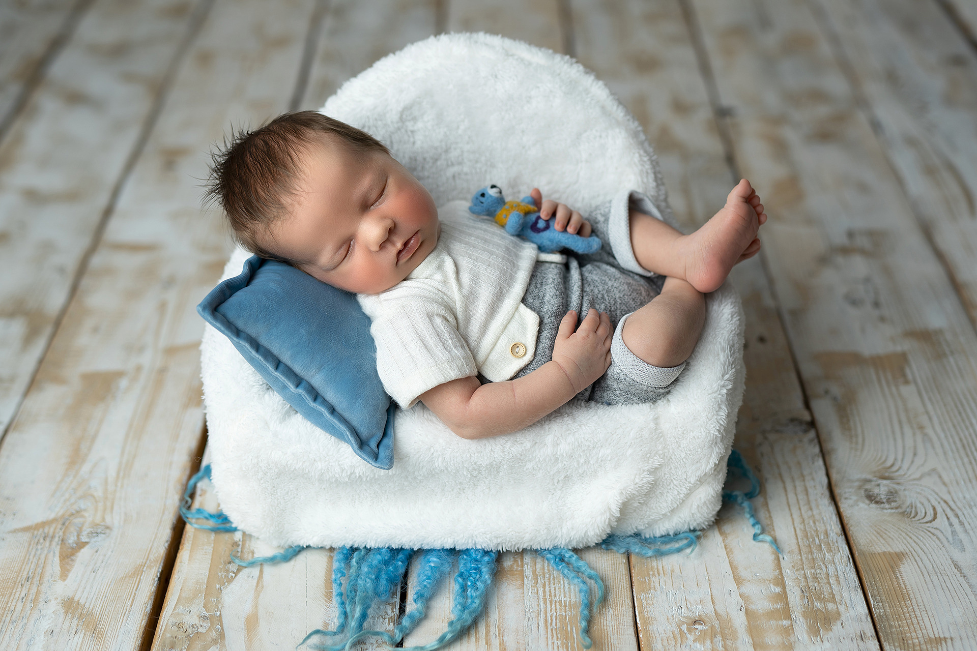 Noworodek śpiący na małym, białym fotelu z pluszowym misiem, na drewnianej podłodze z niebieskimi elementami dekoracyjnymi.