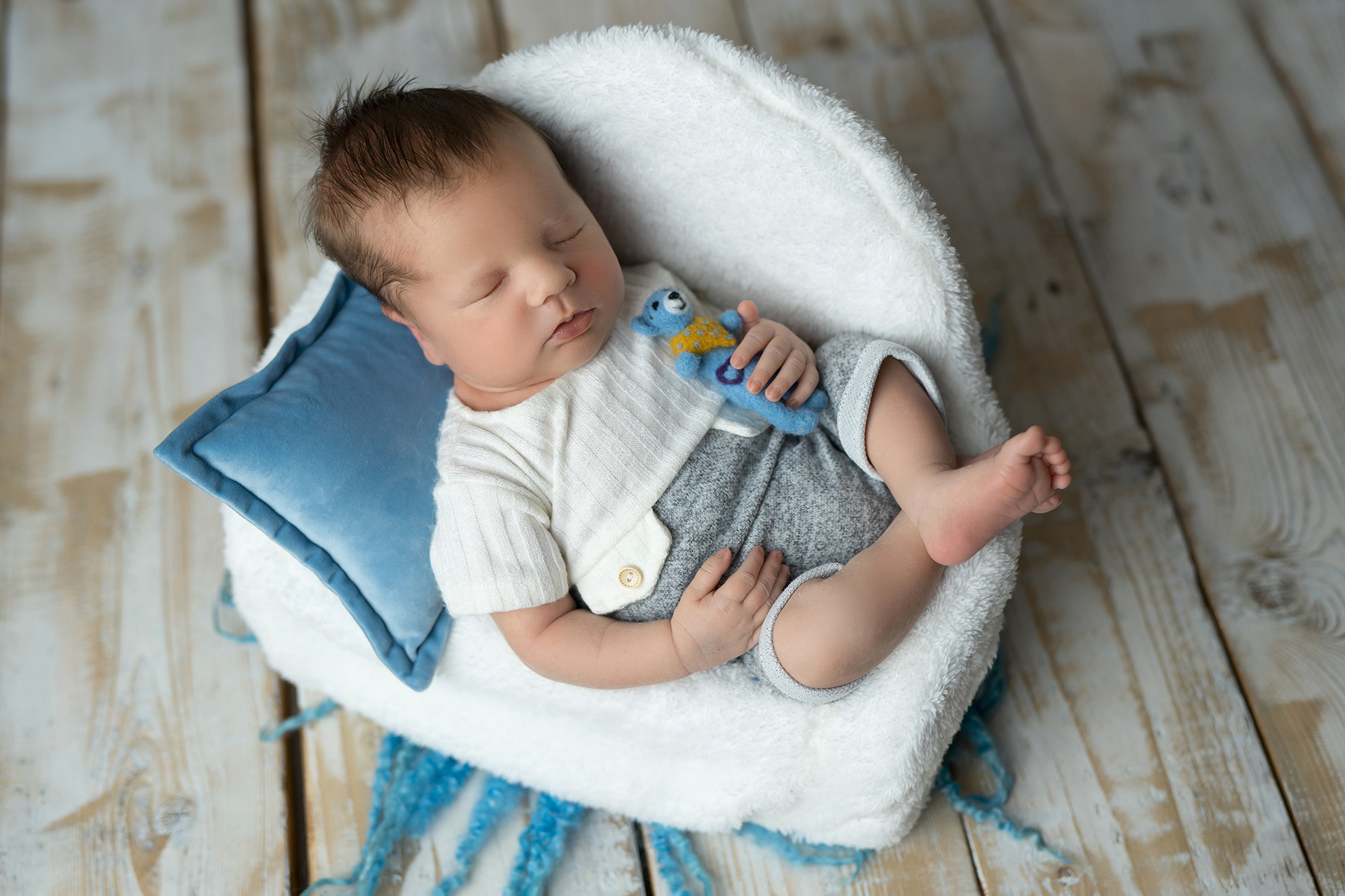 Noworodek śpiący w białym, puszystym gnieździe z niebieską poduszką, trzymający małego, filcowego misia w łapce, na tle drewnianej podłogi.
