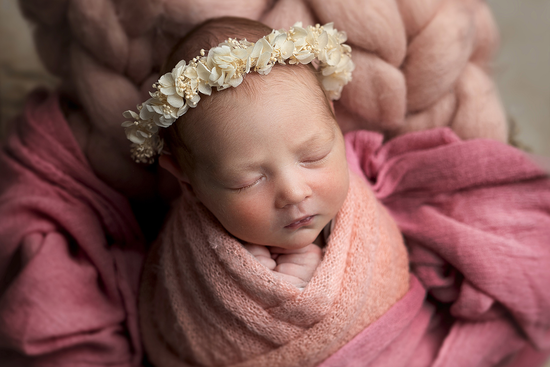Śpiące niemowlę owinięte w różowy kocyk, z rączkami pod brodą i wianuszkiem z jasnych kwiatów na głowie, leżące na tle z grubego, różowego materiału.