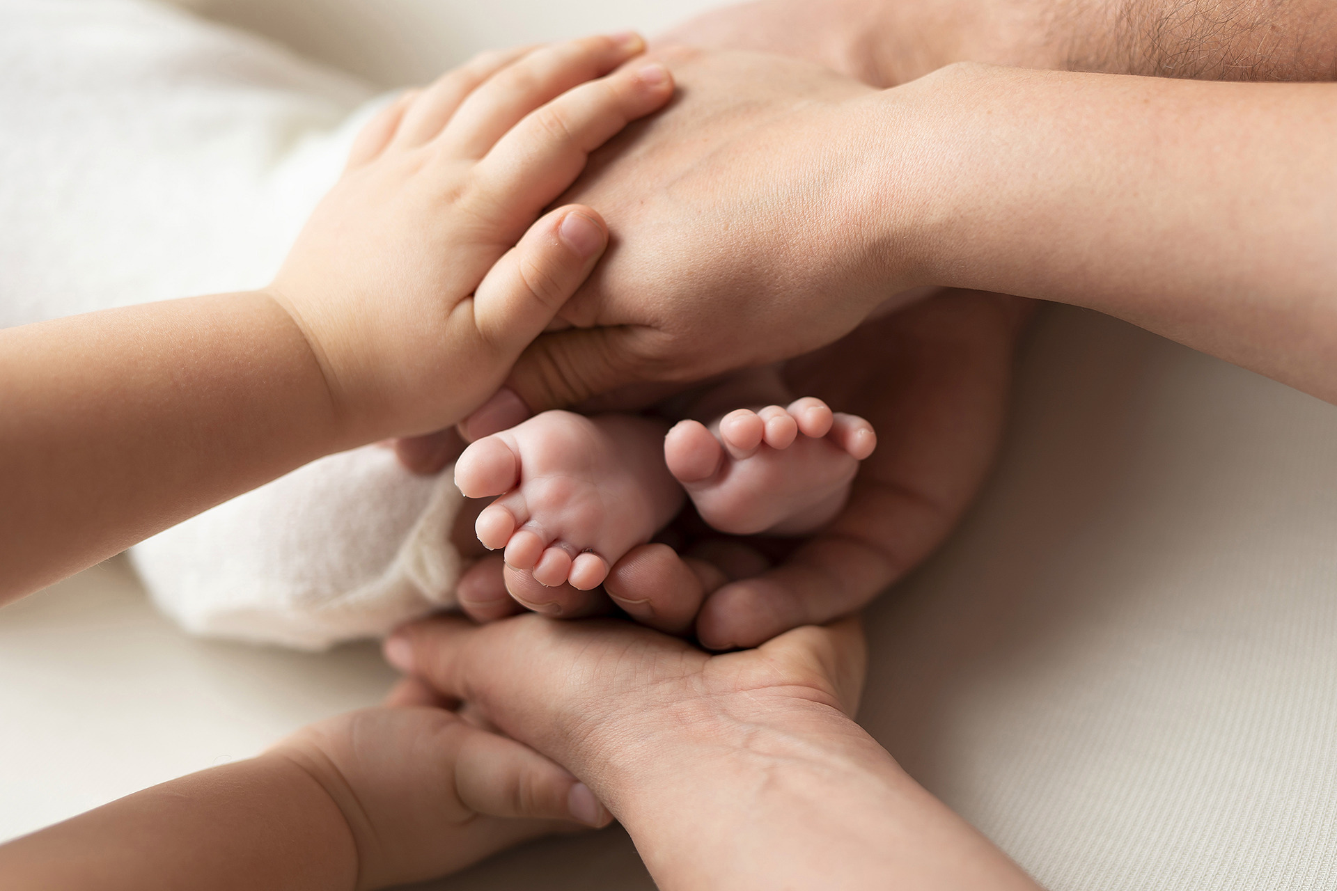 Dłonie rodziców i starszego dziecka otulają stopy noworodka na kremowym tle, delikatne oświetlenie.