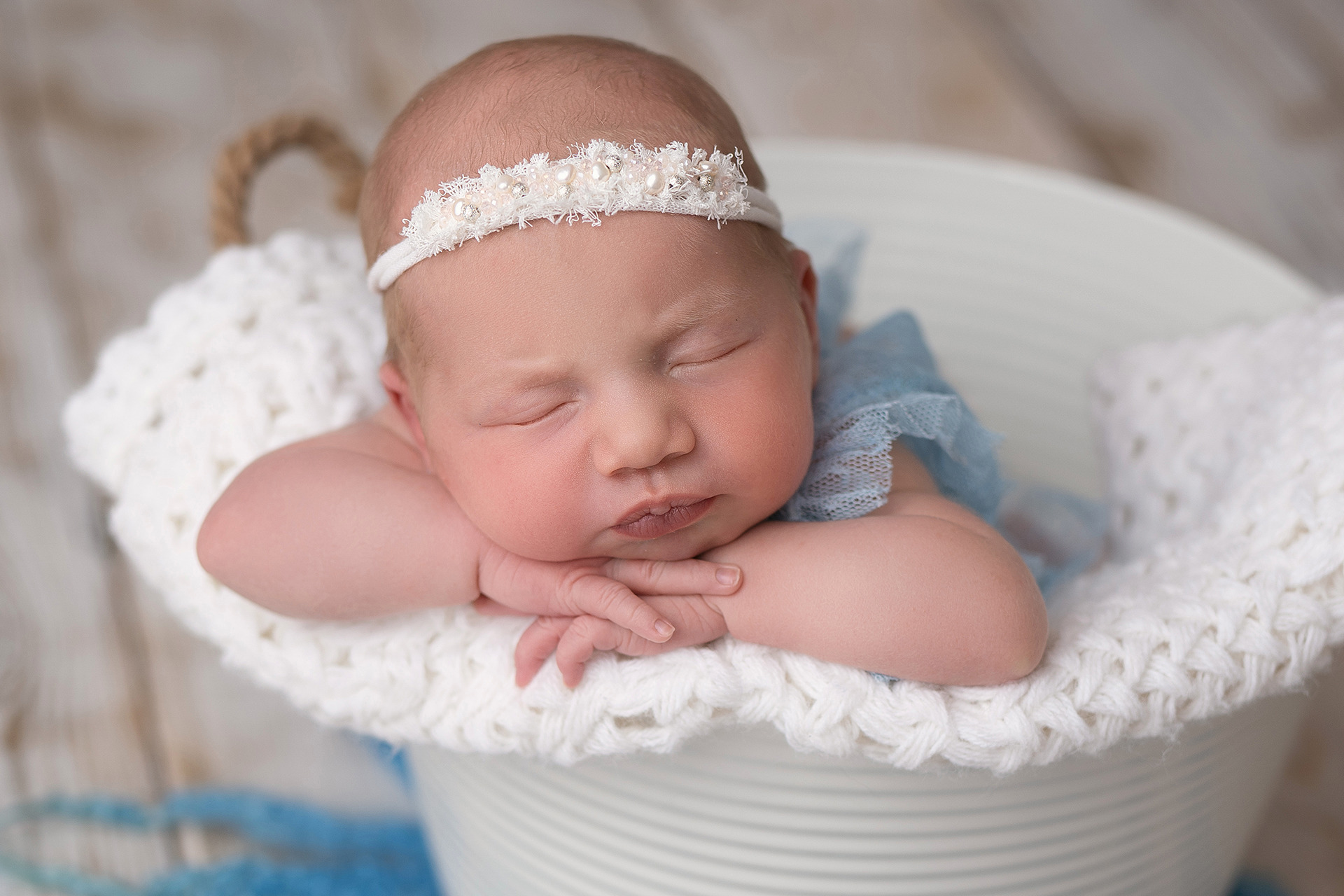 Noworodek w białym koszyku, z koronkową opaską z perełkami, śpiący na dzierganym kocyku z niebieskimi akcentami.