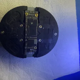 Płyta główna smartfona umieszczona w okrągłym uchwycie, widoczne elementy elektroniczne i złącza, całość na tle białej serwetki i niebieskiego podłoża.