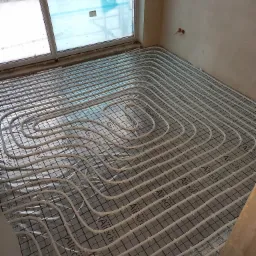 Rozłożone rury ogrzewania podłogowego na folii z rastrem w surowym wnętrzu, widoczne okno z folią ochronną.