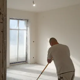 Mężczyzna w białych spodniach roboczych maluje wałkiem podłogę w jasnym, odnawianym pomieszczeniu z oknem; widoczna surowa rama drzwi i wisząca żarówka.