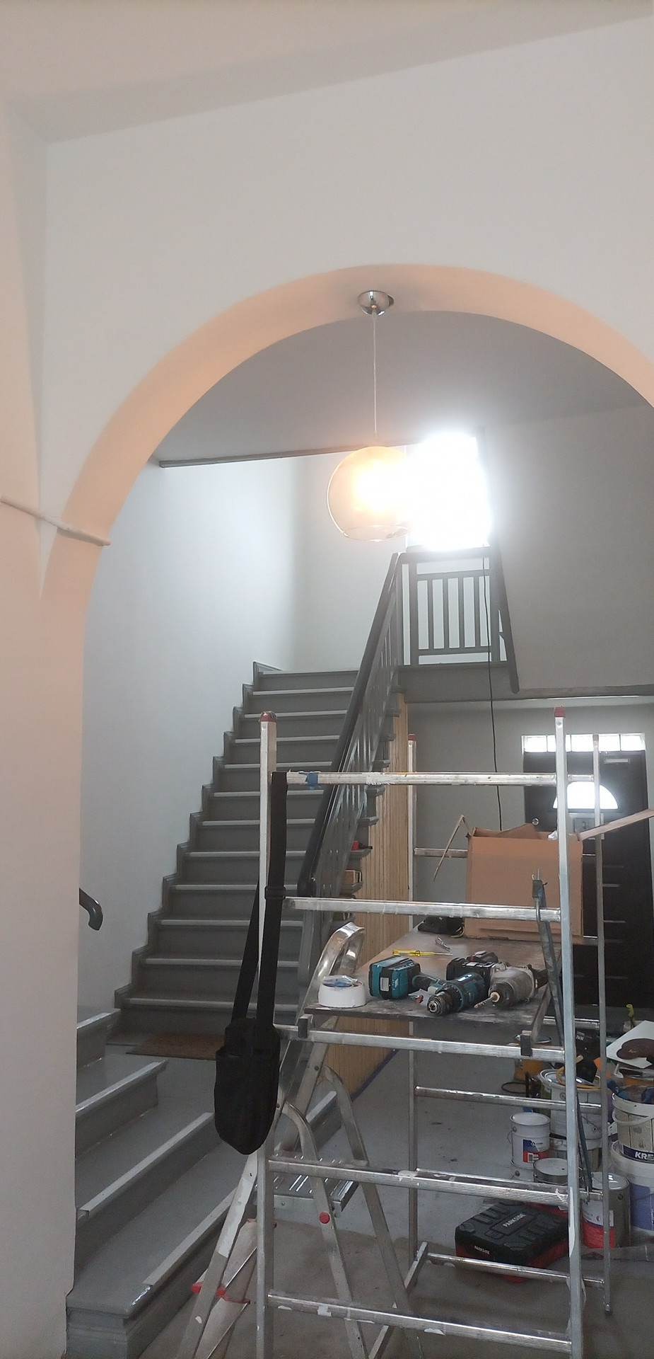 Klatka schodowa w trakcie malowania, widoczne rusztowanie z narzędziami i farbami, świeżo pomalowane schody i ściany w odcieniach szarości i bieli, lampa wisząca i łukowe przejście.
