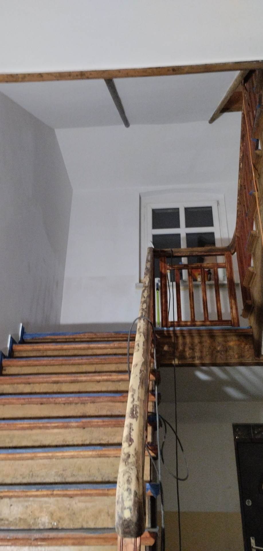 Schody drewniane zabezpieczone taśmą malarską przed malowaniem ścian, widoczne ślady użytkowania i odchodząca farba z poręczy.