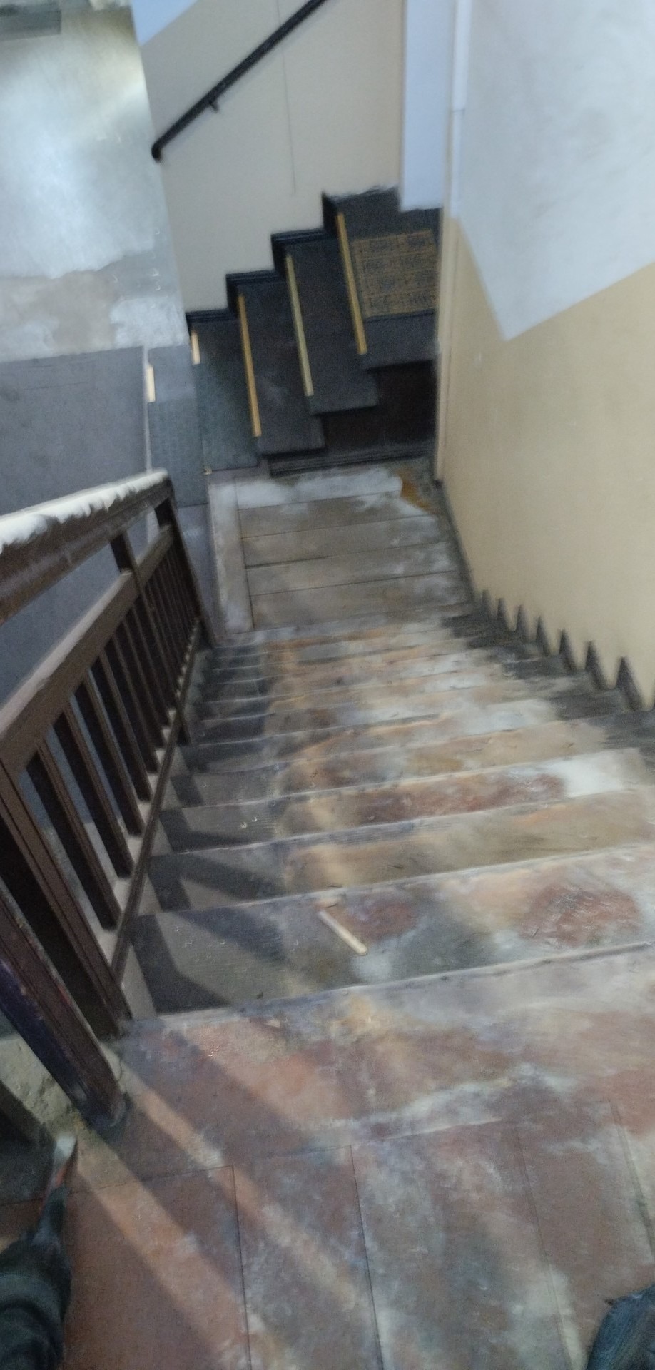 Klatka schodowa w trakcie remontu; widoczne stare schody z pozostałościami farby, drewniana balustrada i fragment ściany z widocznymi śladami napraw.