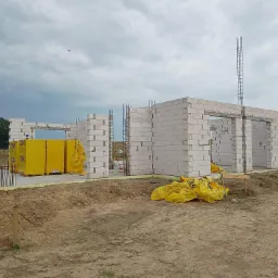 Wznoszenie ścian domu z białych bloczków Ytong, widoczne zbrojenia pionowe, żółte opakowania bloczków i folia ochronna, teren budowy z ziemią.