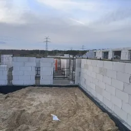 Wznoszenie ścian parterowego domu z białych bloczków, widoczne stalowe pręty zbrojeniowe, teren wokół zasypany piaskiem, w tle słupy wysokiego napięcia.