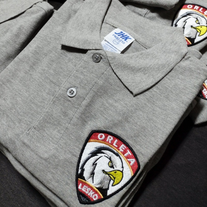 Stos szarych koszulek polo z haftowanym herbem 'Orlęta Lesko' na piersi, widoczna metka JHK PKID 210.