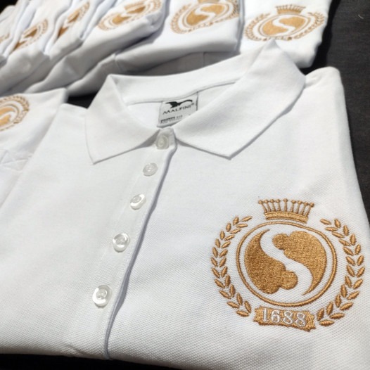 Biała koszulka polo z wyhaftowanym złotym herbem z koroną i datą 1688, leżąca na stosie innych koszulek z tym samym wzorem.